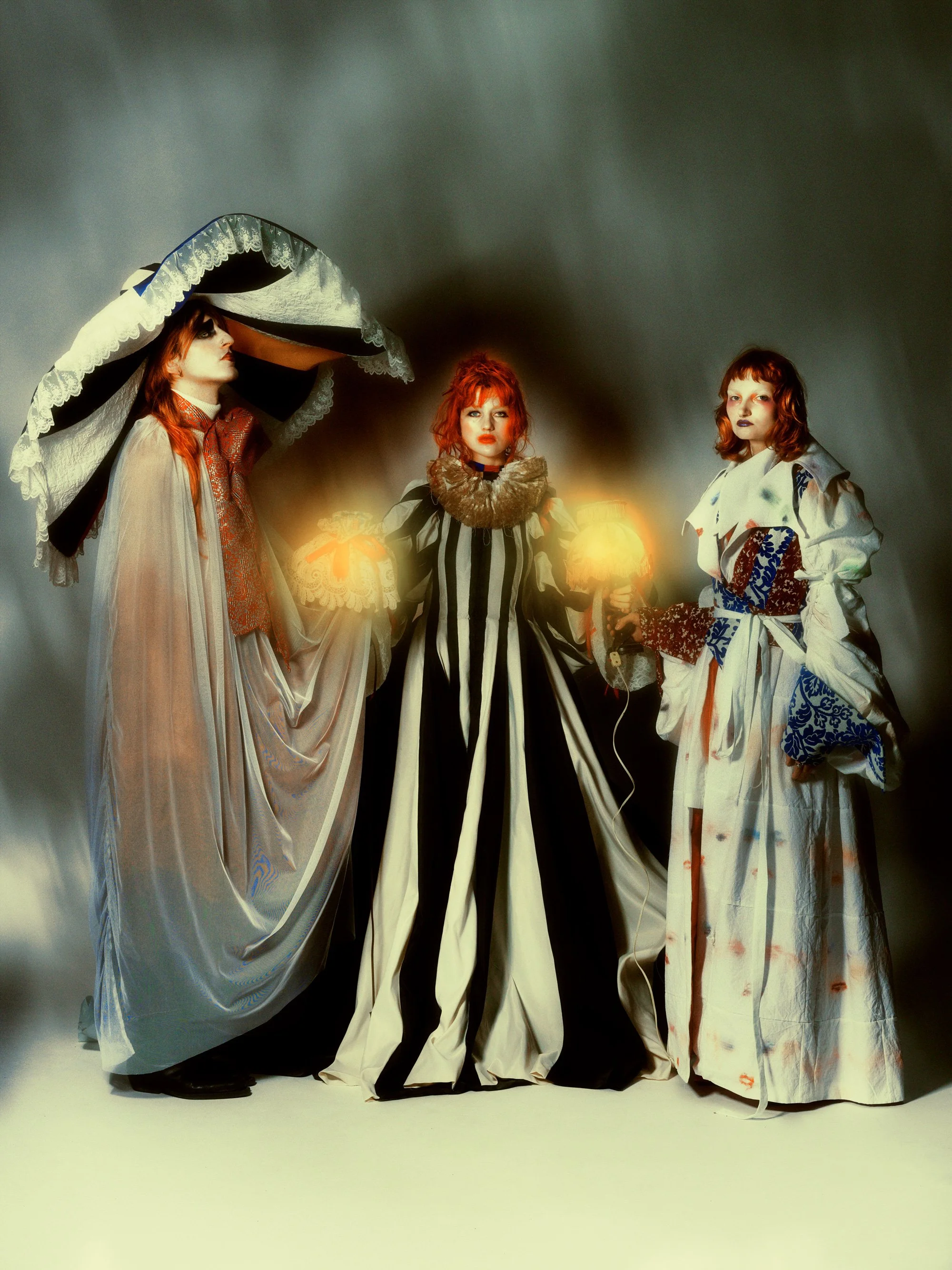 Trois femmes vêtues de robes longues et élégantes, tenant des lampes ou des objets lumineux, posant contre un fond gris sombre.