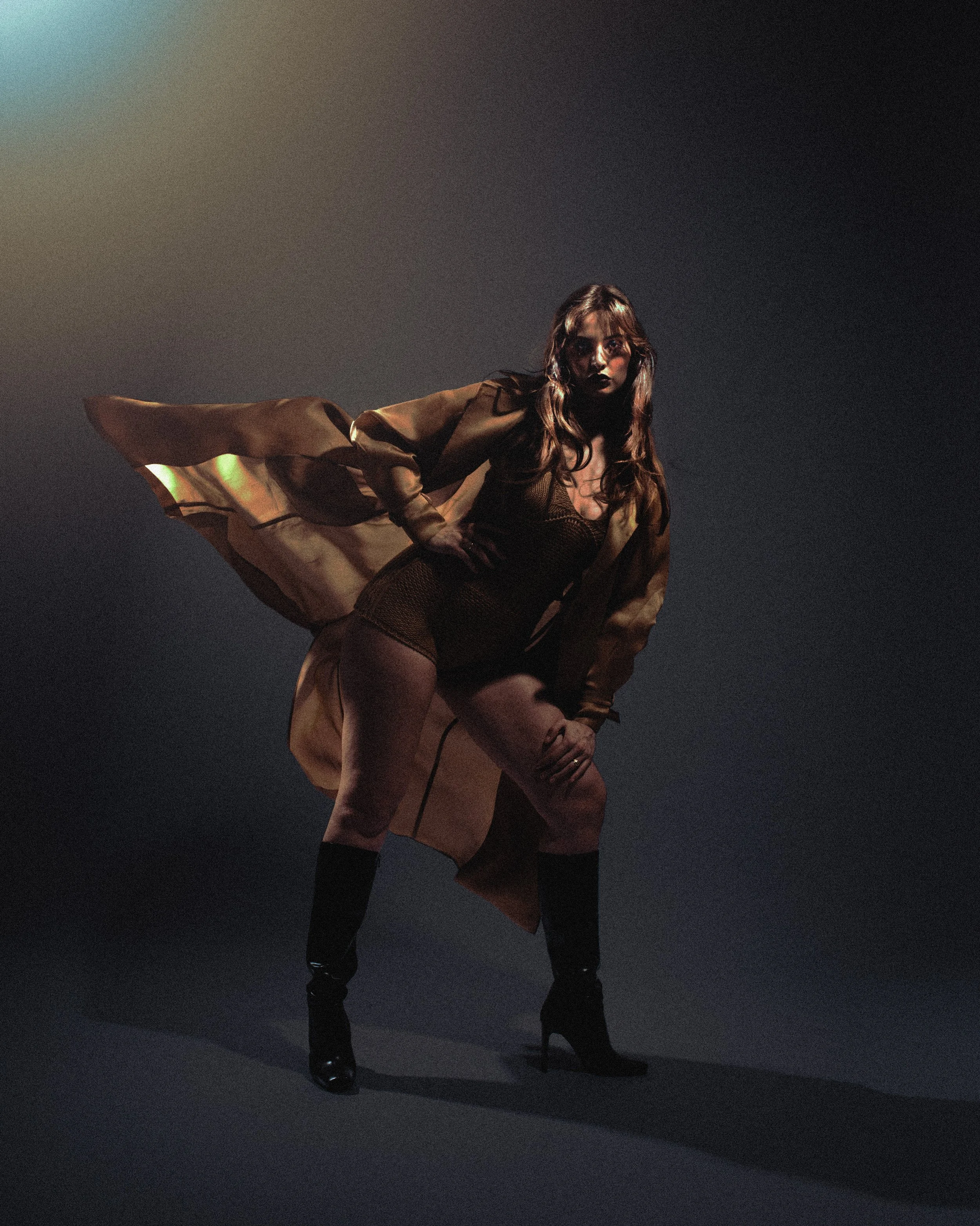 Une femme pose dans une lumière sombre, portant un manteau beige, un body noir, des chaussettes hautes en cuir et des talons hauts noirs.