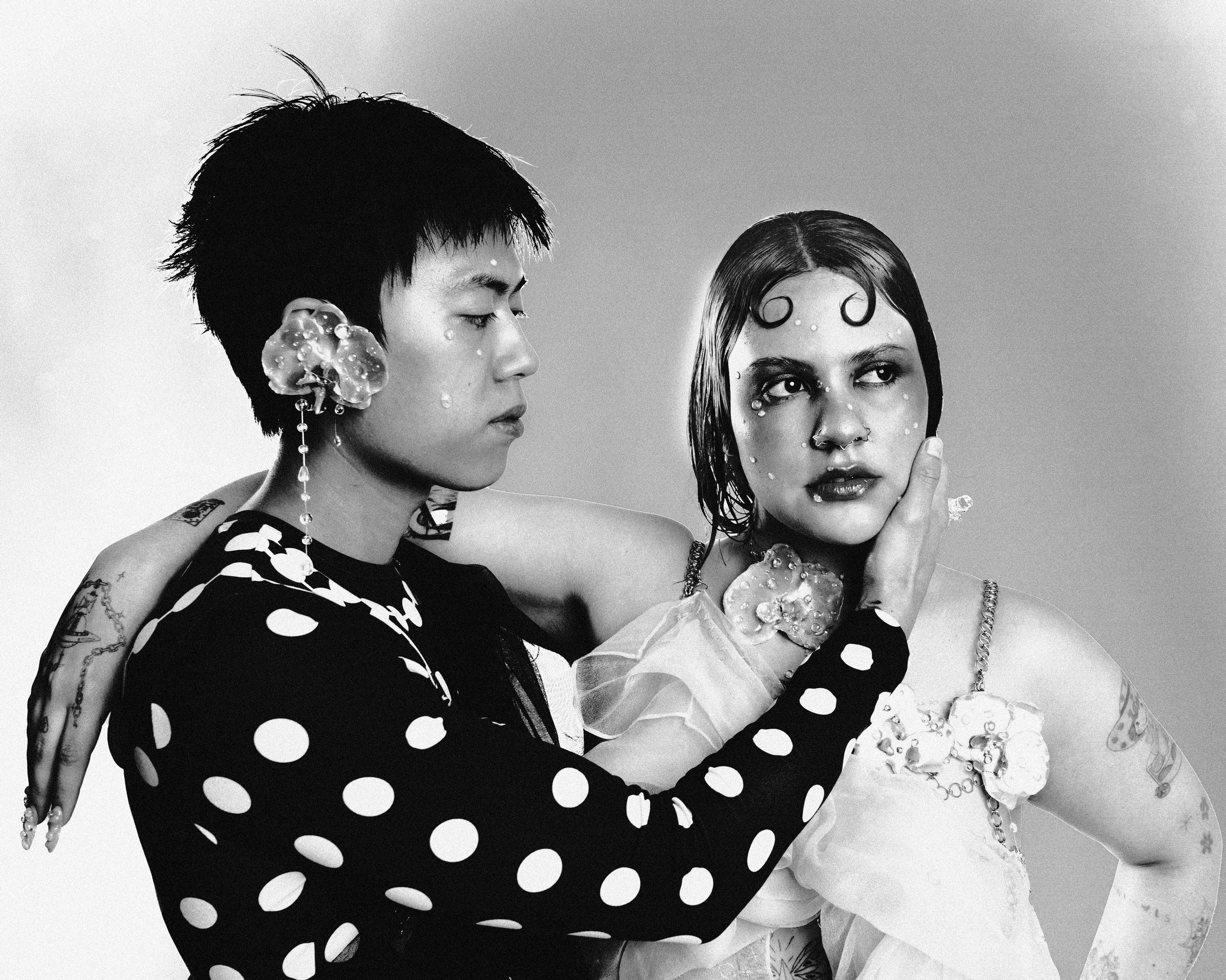 Deux personnes posent ensemble, une à gauche et l'autre à droite, dans un style artistique et avant-gardiste, avec des accessoires et un maquillage distinctifs, en noir et blanc.