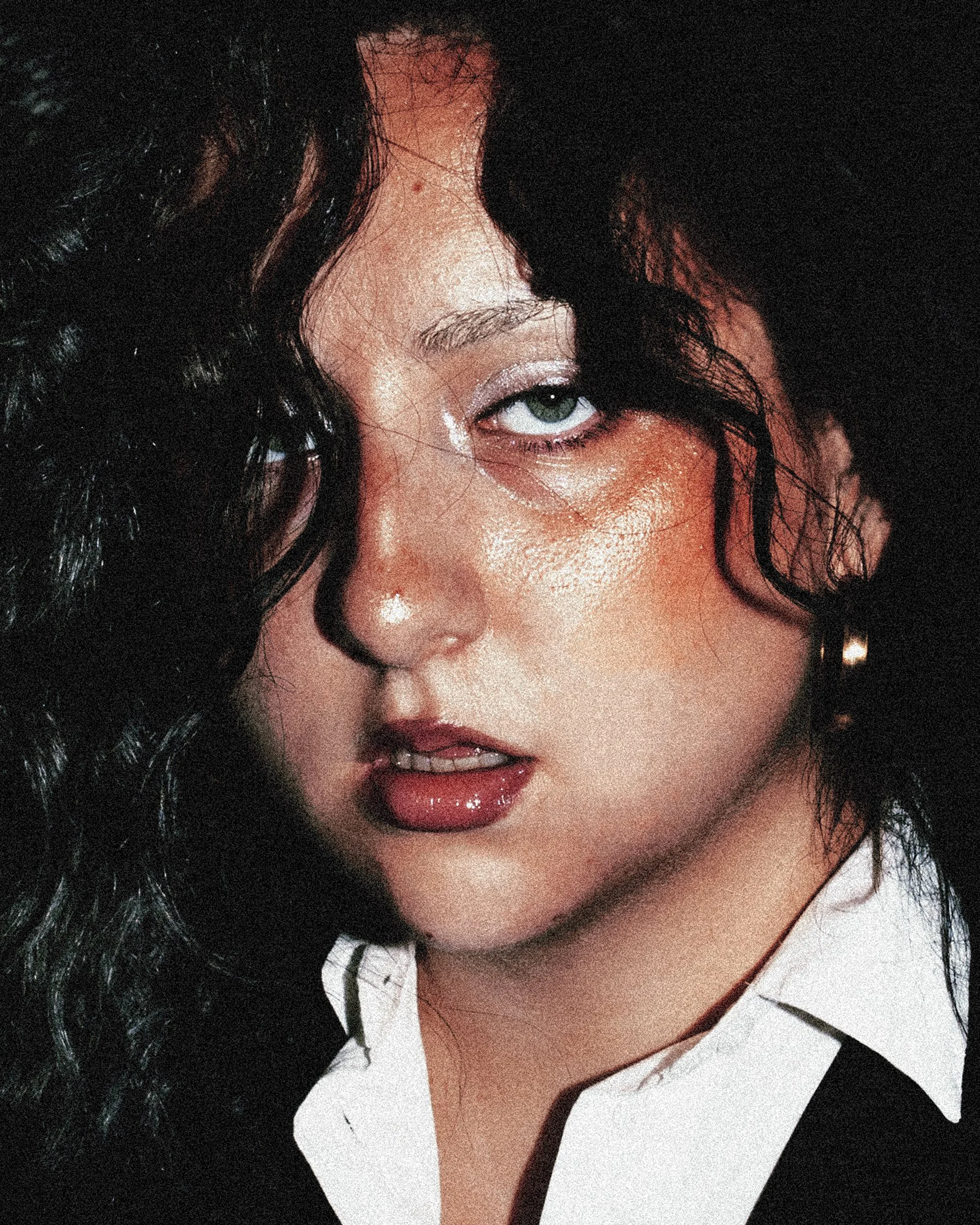 Premier plan d'une femme avec des cheveux noirs bouclés, maquillage brillant, lèvres pulpeuses, portant une chemise blanche et un bouclé d'oreille.