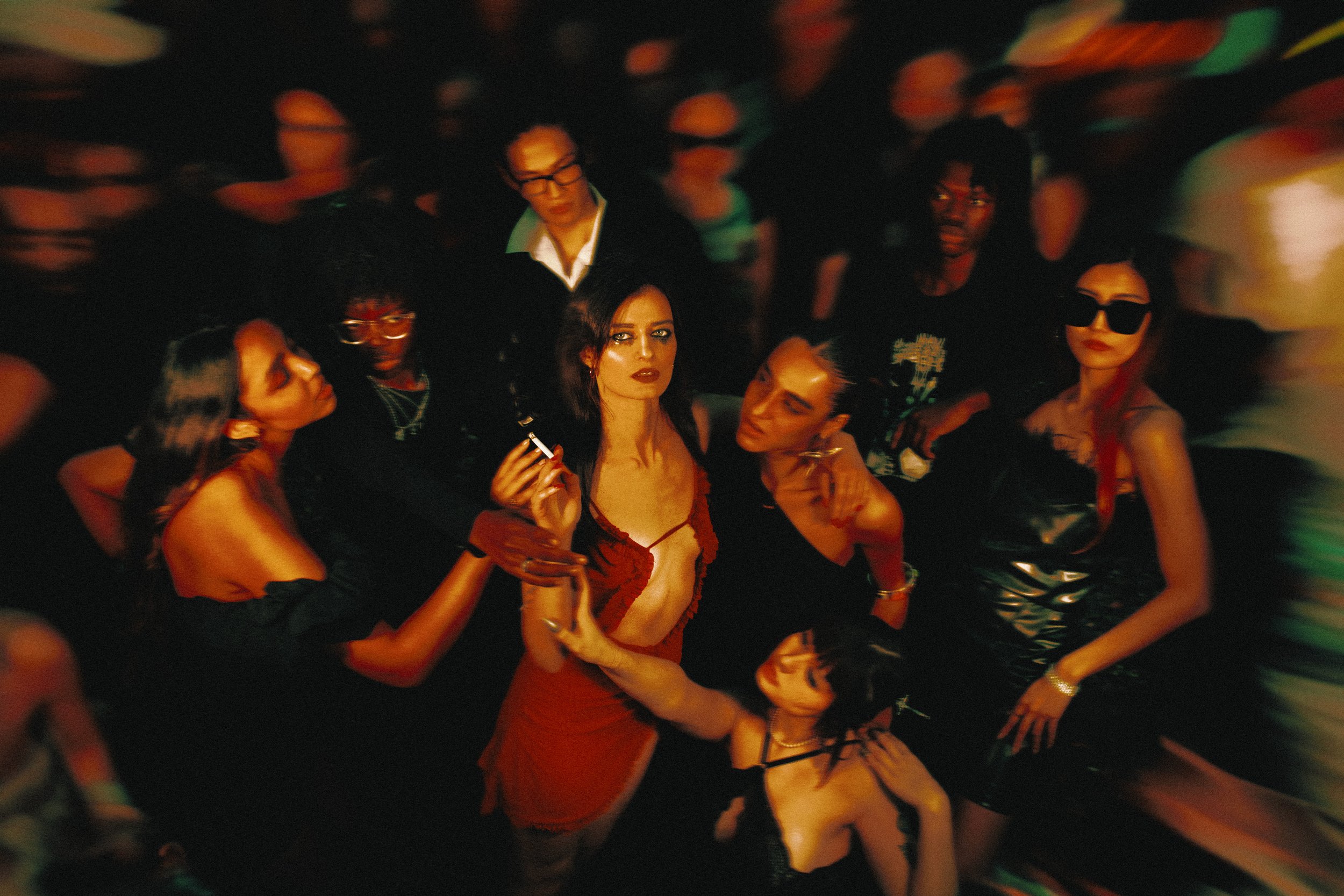 Un groupe de personnes dans une ambiance sombre et colorée, semblant être dans une fête ou un club, avec des individus portant des vêtements élégants et sophistiqués.