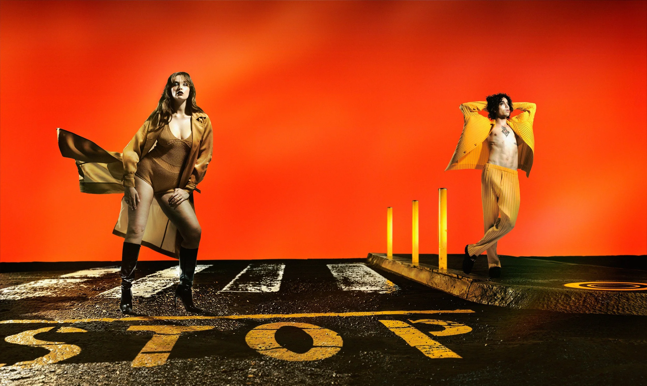 Deux personnes posant sur un sol de stationnement avec un fond orange. La personne à gauche porte un manteau ocre, un maillot moulant et des bottes à talons, alors que la personne à droite, pieds nus, porte une veste de couleur jaune et un pantalon rayé, tout en étant dénudée sur le torse.