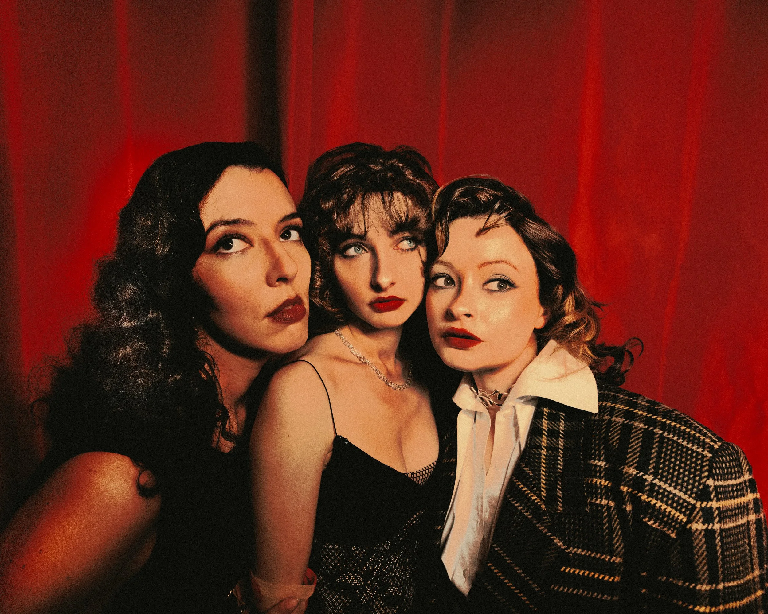 Trois femmes posent devant un rideau rouge, affichant un style rétro et sophistiqué.