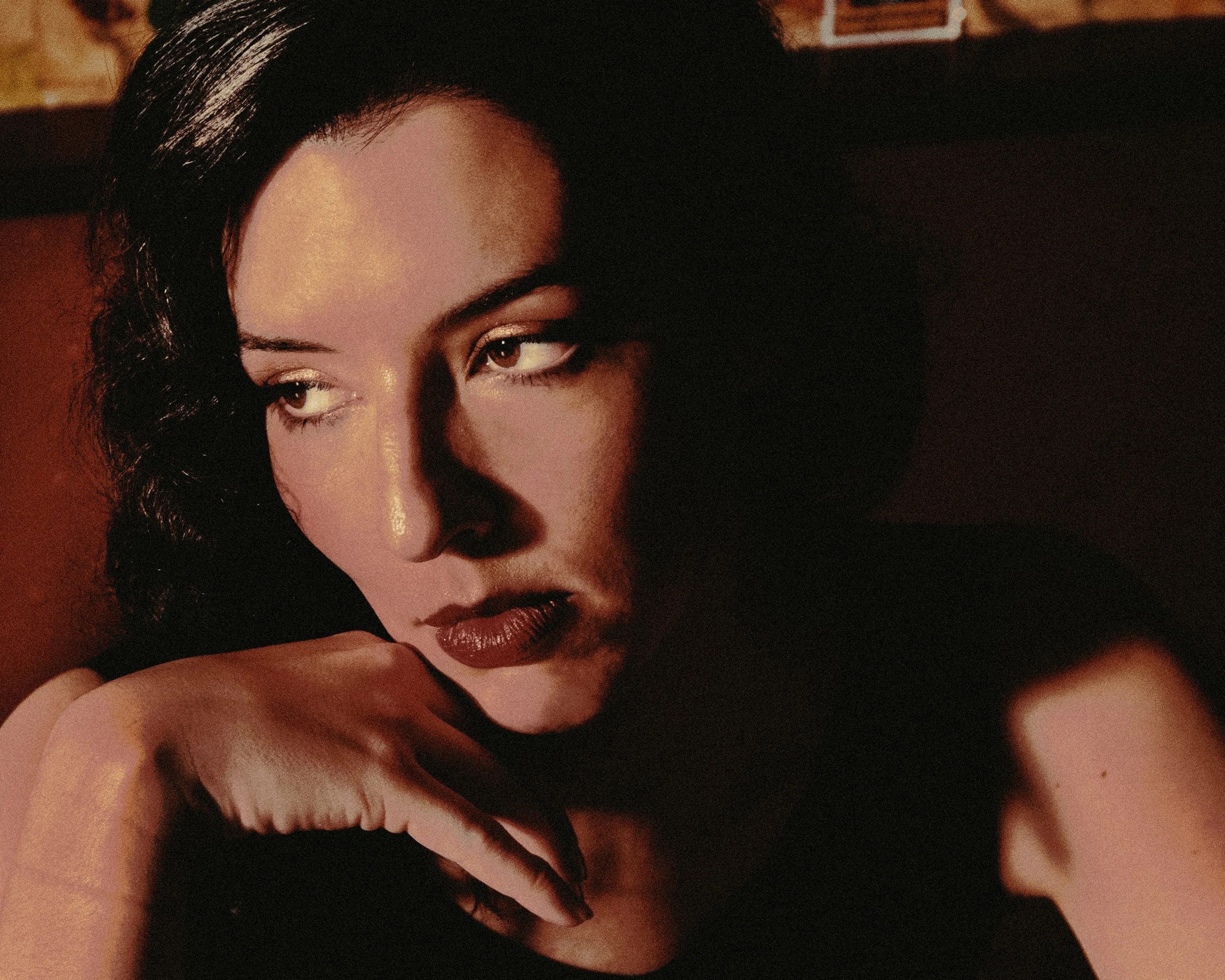 Portrait d'une femme aux cheveux noirs, regardant vers la gauche, avec une expression sérieuse, dans un éclairage chaud et sombre.