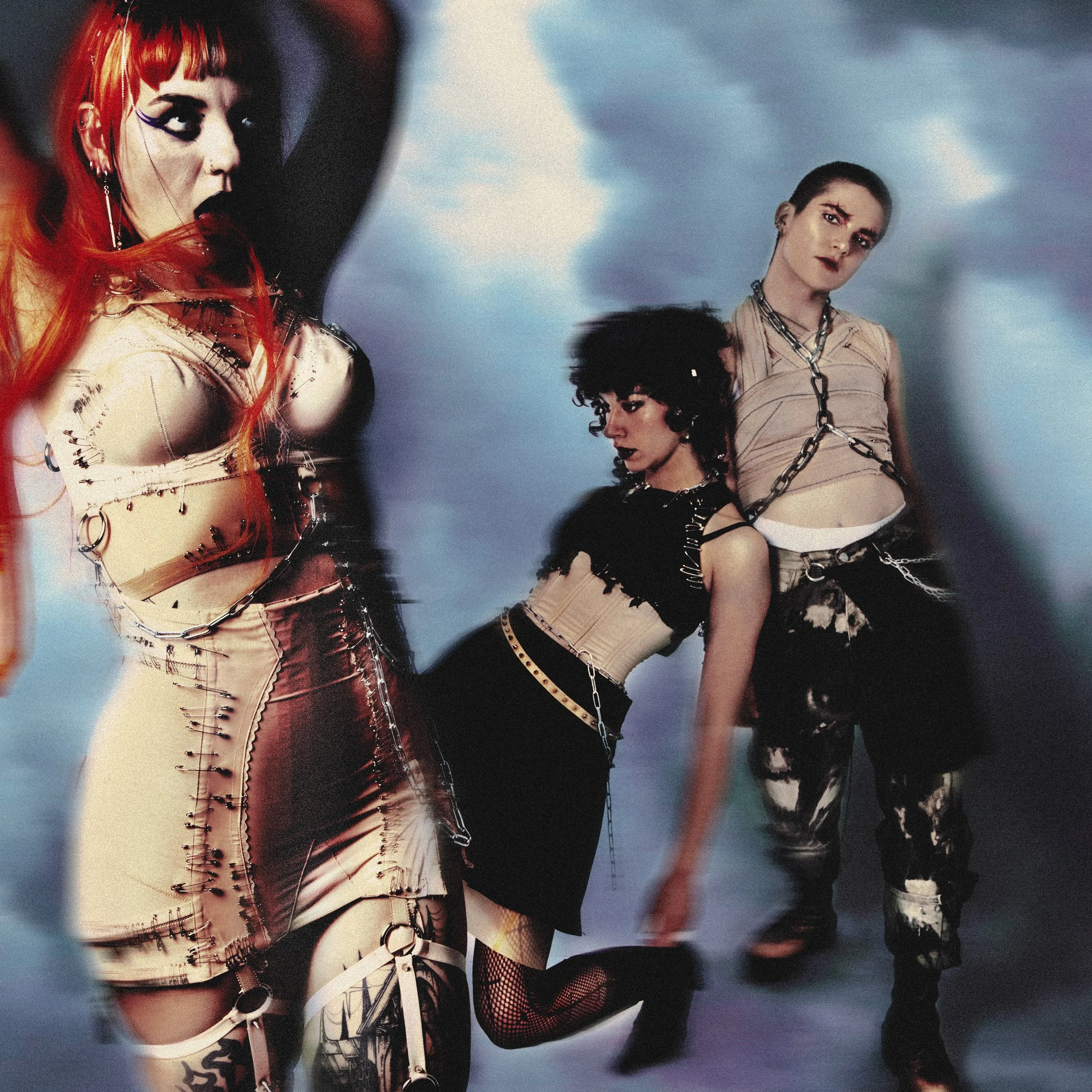 Trois femmes posent dans un style punk ou gothique avec des vêtements en latex et des accessoires en chaînes, sous un ciel grisâtre.