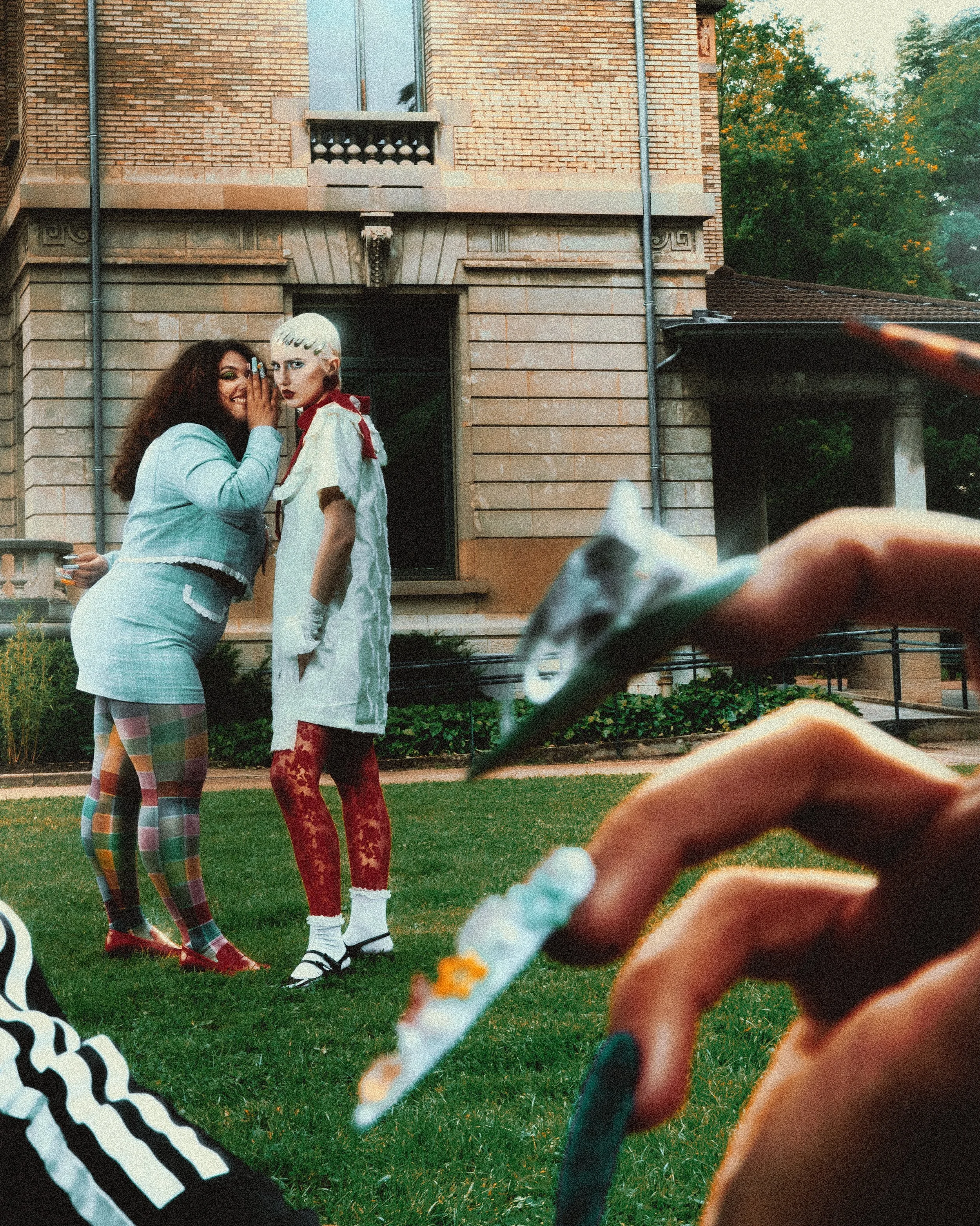 Deux femmes posant de manière amusante devant une maison en briques, une tenant une cigarette, une autre étant vêtue de façon colorée avec un style audacieux. La photo est prise de près, avec une main en premier plan tenant une cigarette.