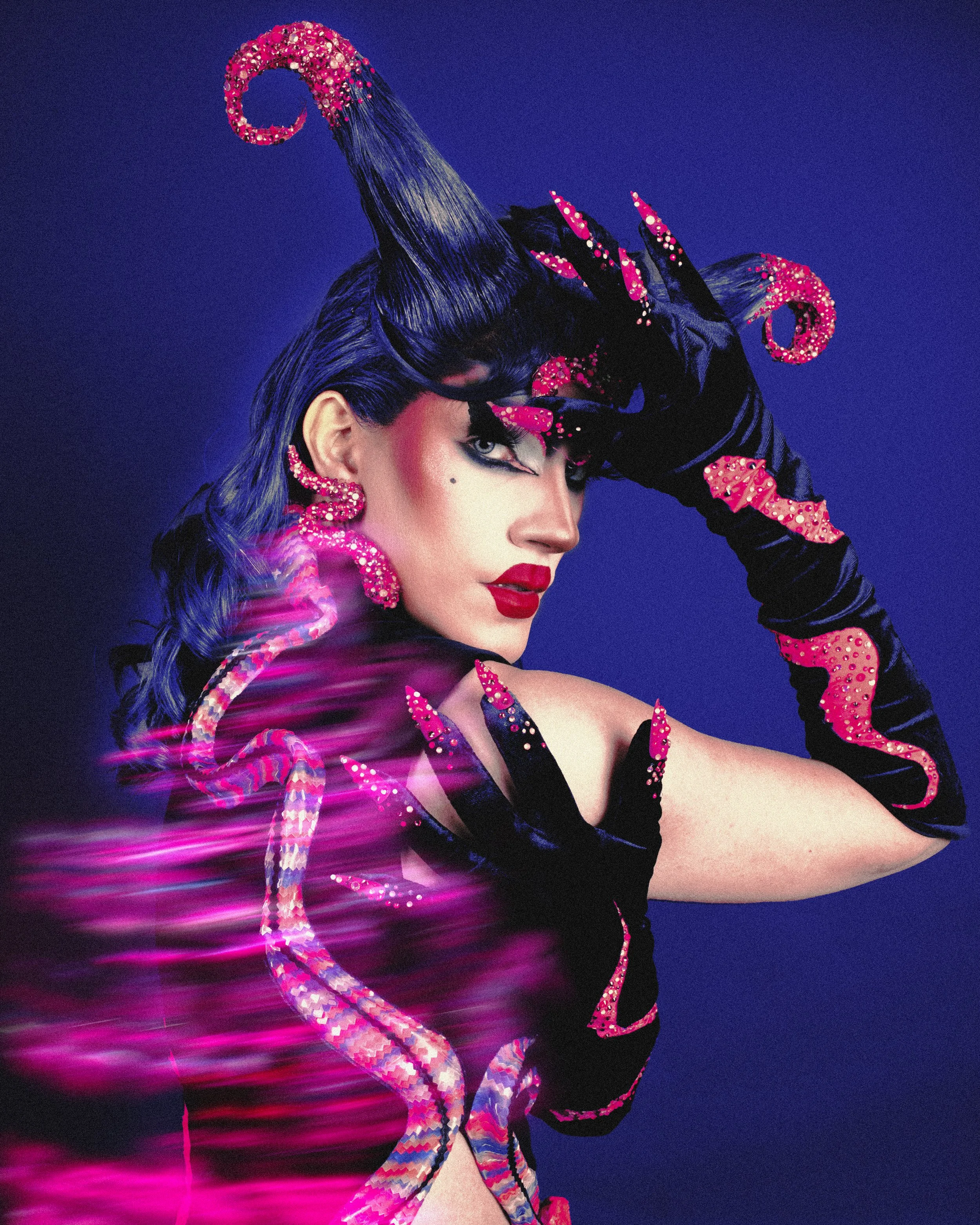 Une femme avec cheveux bleus et maquillage audacieux, portant des gants et des accessoires décorés de motifs de serpents roses et à paillettes, posant contre un fond violet.