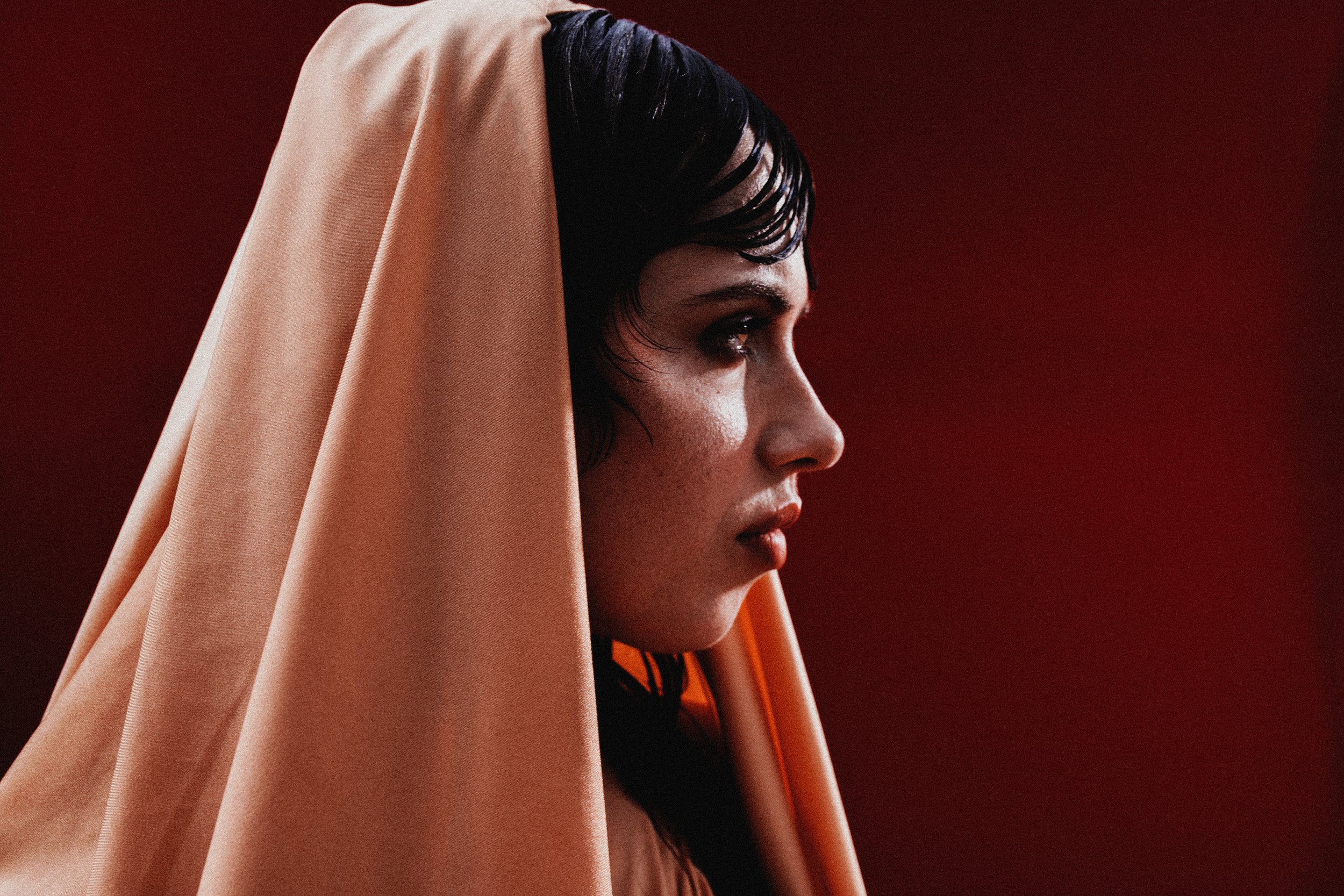 Portrait d'une femme avec une coiffure courte noire, portant un voile beige, regardant vers la droite, fond rouge