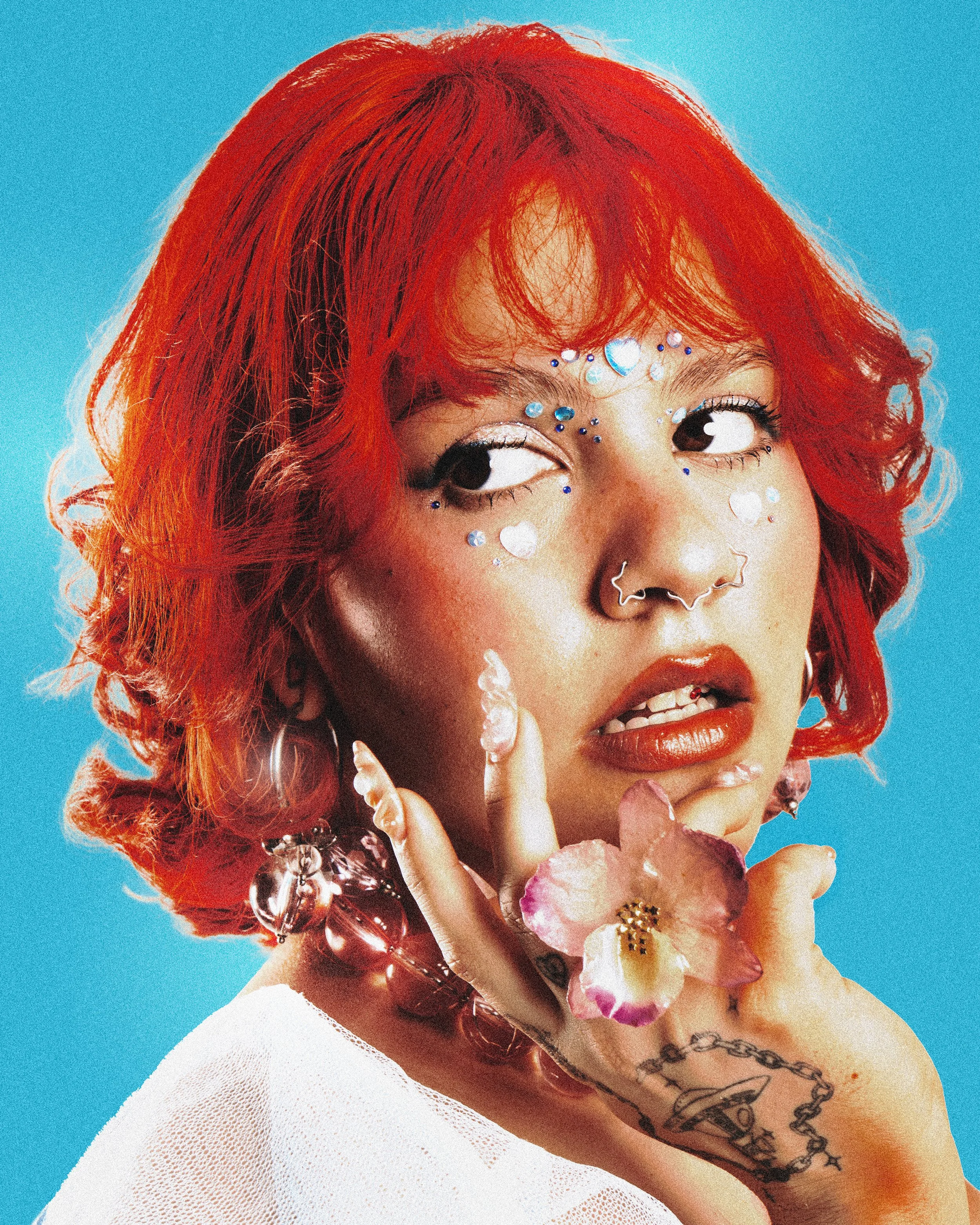 Jeune femme avec cheveux courts et rouges, portant des bijoux, des décorations de visage et un maquillage coloré, tenant une fleur près du visage, avec un fond bleu.