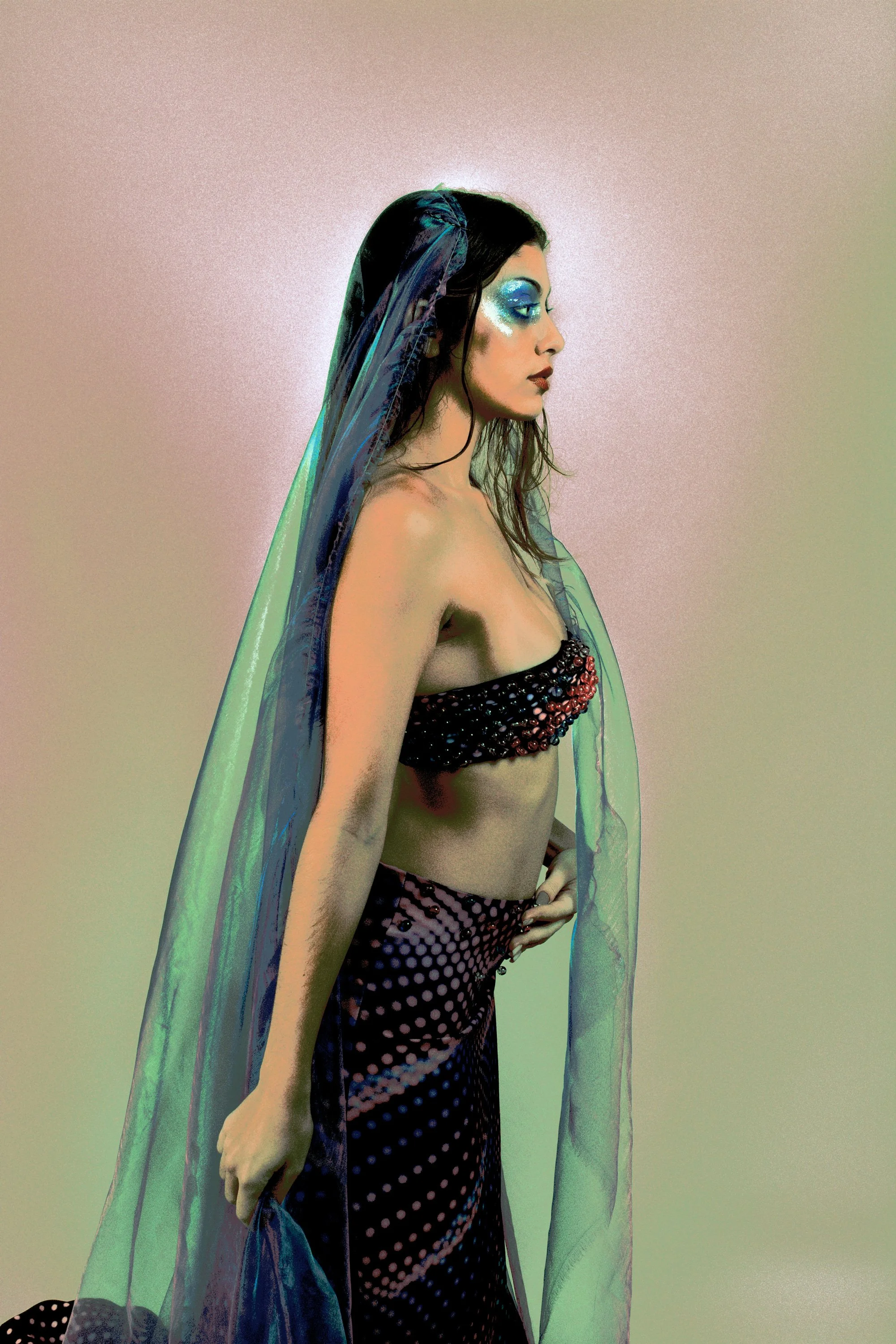 Femme portant un voile transparent, vêtue d'un haut et d'une jupe à motifs, maquillée avec de la crème glacée bleu sur les yeux, pose contre un fond neutre.