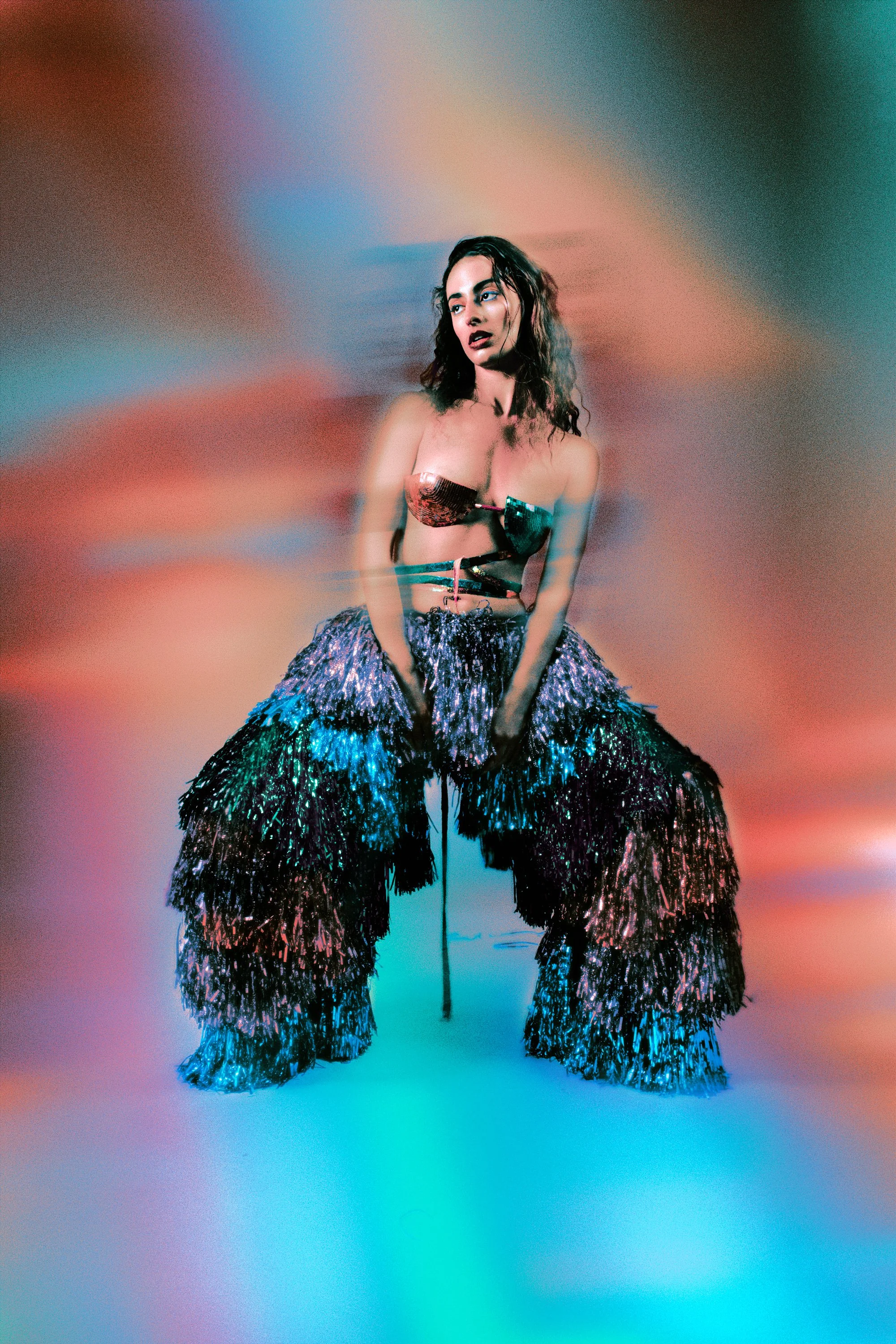 Une femme portant un haut en miroir et un pantalon en franges iridescentes, assise ou pliée en genoux devant un fond flou aux couleurs pastel