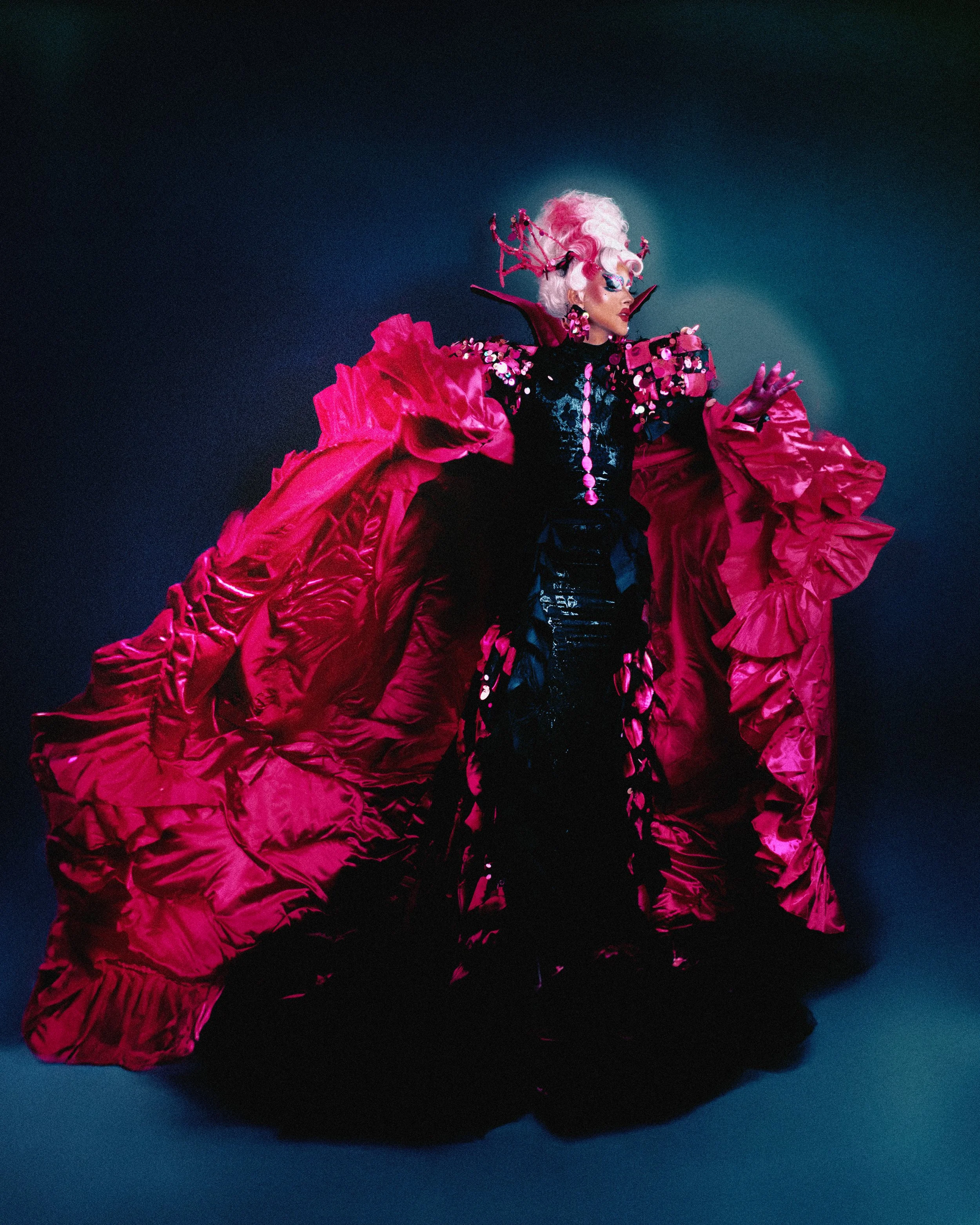 Une personne habillée en costume extravagant avec une robe rouge à volants, coiffée d'une perruque blanche ondulée et de cornes, portant des accessoires roses, sur fond sombre.