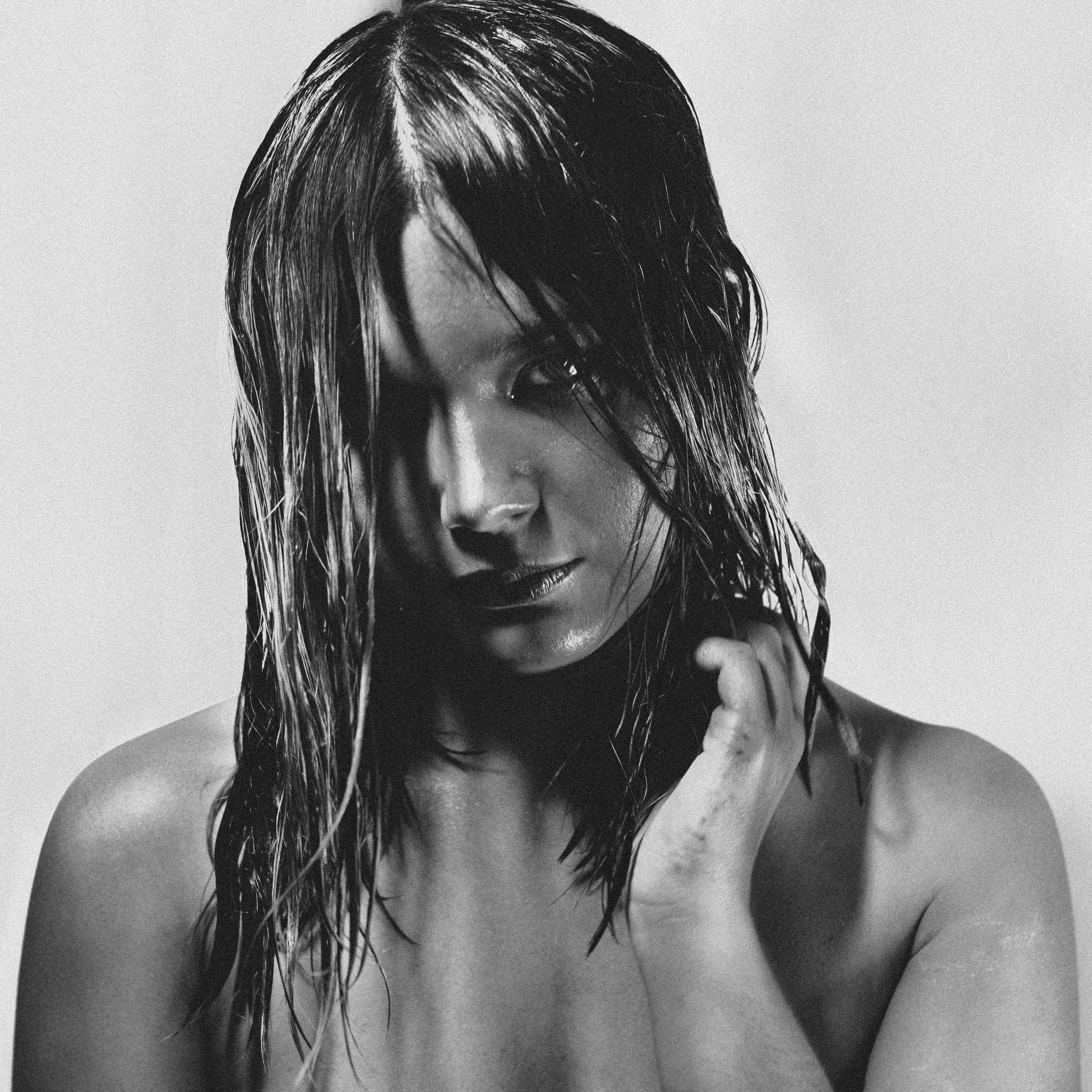 Une femme avec les cheveux mouillés, en noir et blanc, regardant vers l'objectif avec une expression intense, ses épaules nues visibles.