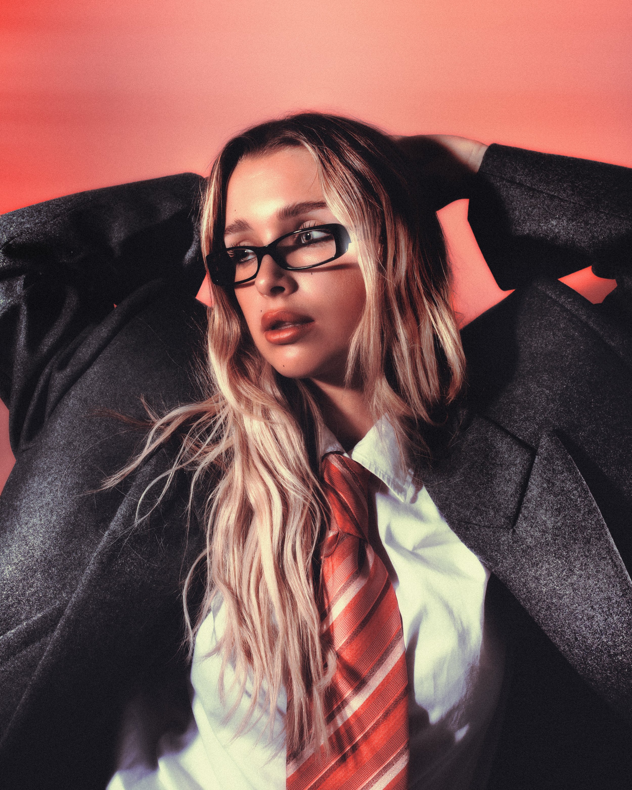 Jeune femme portant des lunettes noires, une chemise blanche, une cravate rayée orange et un manteau gris foncé, avec les cheveux longs et ondulés, contre un fond rose.