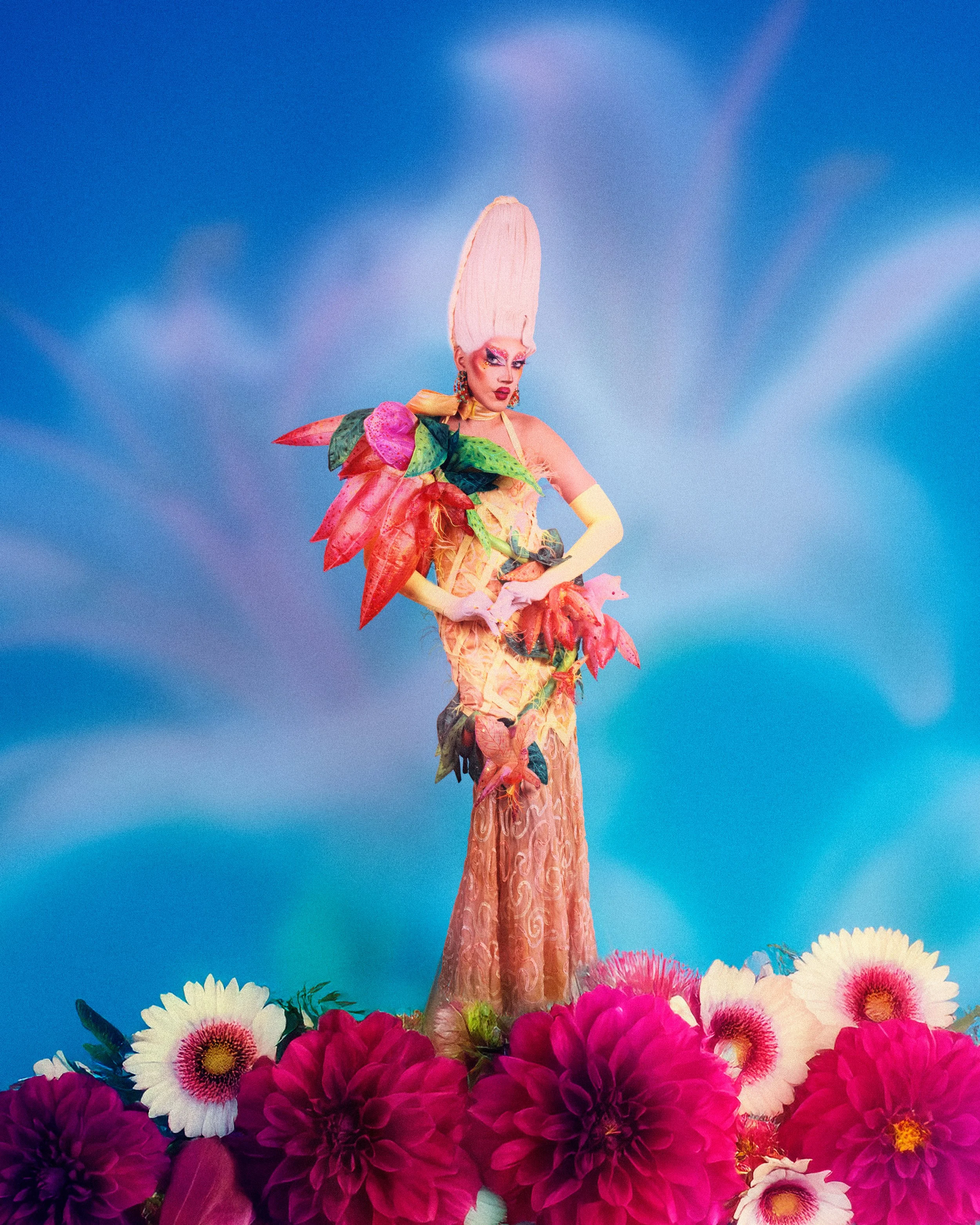 Une personne vêtue d'une robe florale colorée, portant un grand chapeau rose, entourée de fleurs dans un décor coloré avec un ciel bleu en arrière-plan.