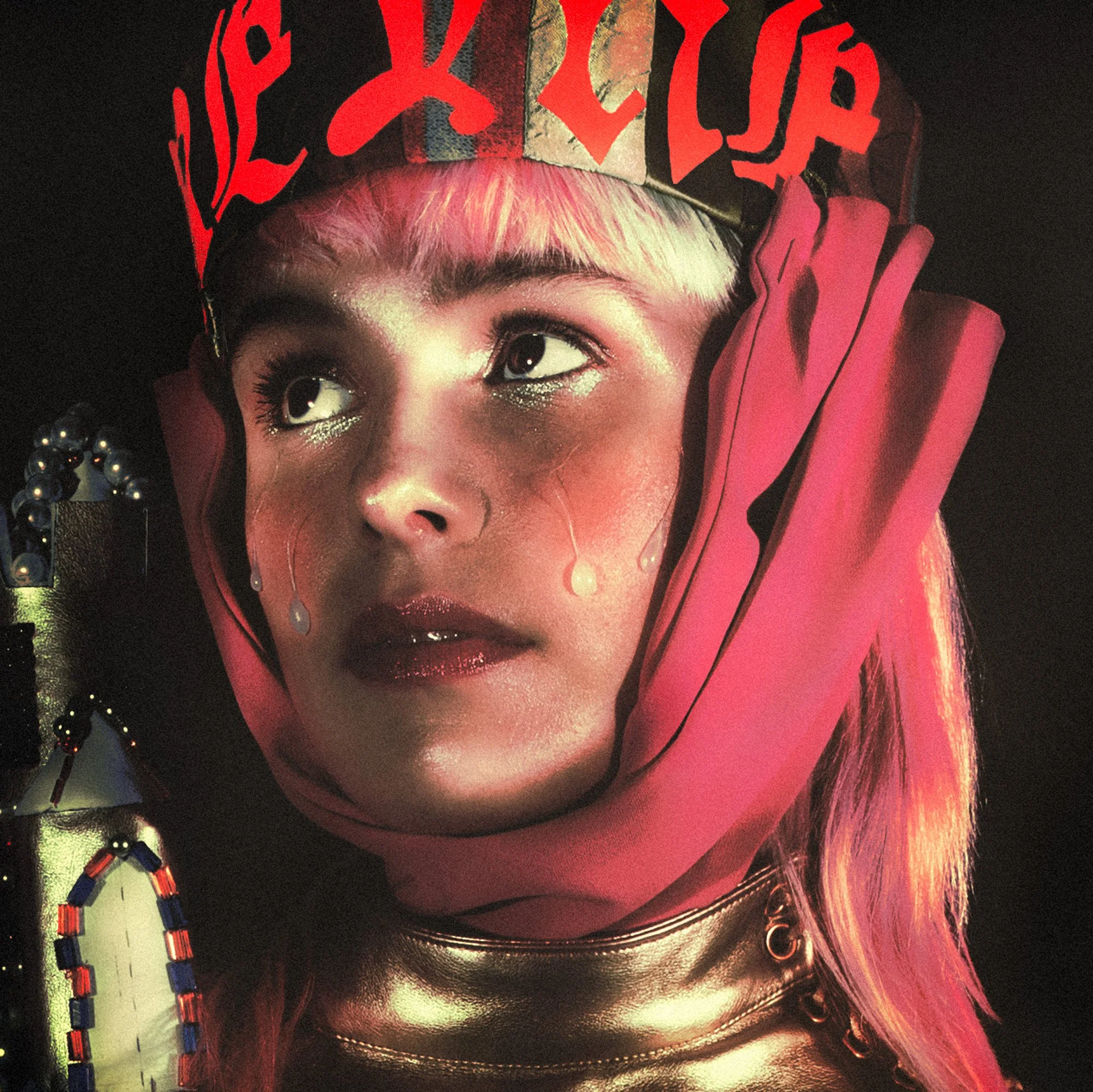 Une jeune femme avec un maquillage brillant, portant un casque avec des motifs rouges et une capuche rose. Elle a les cheveux courts et blonds avec des reflets rose, et porte un collier en métal.