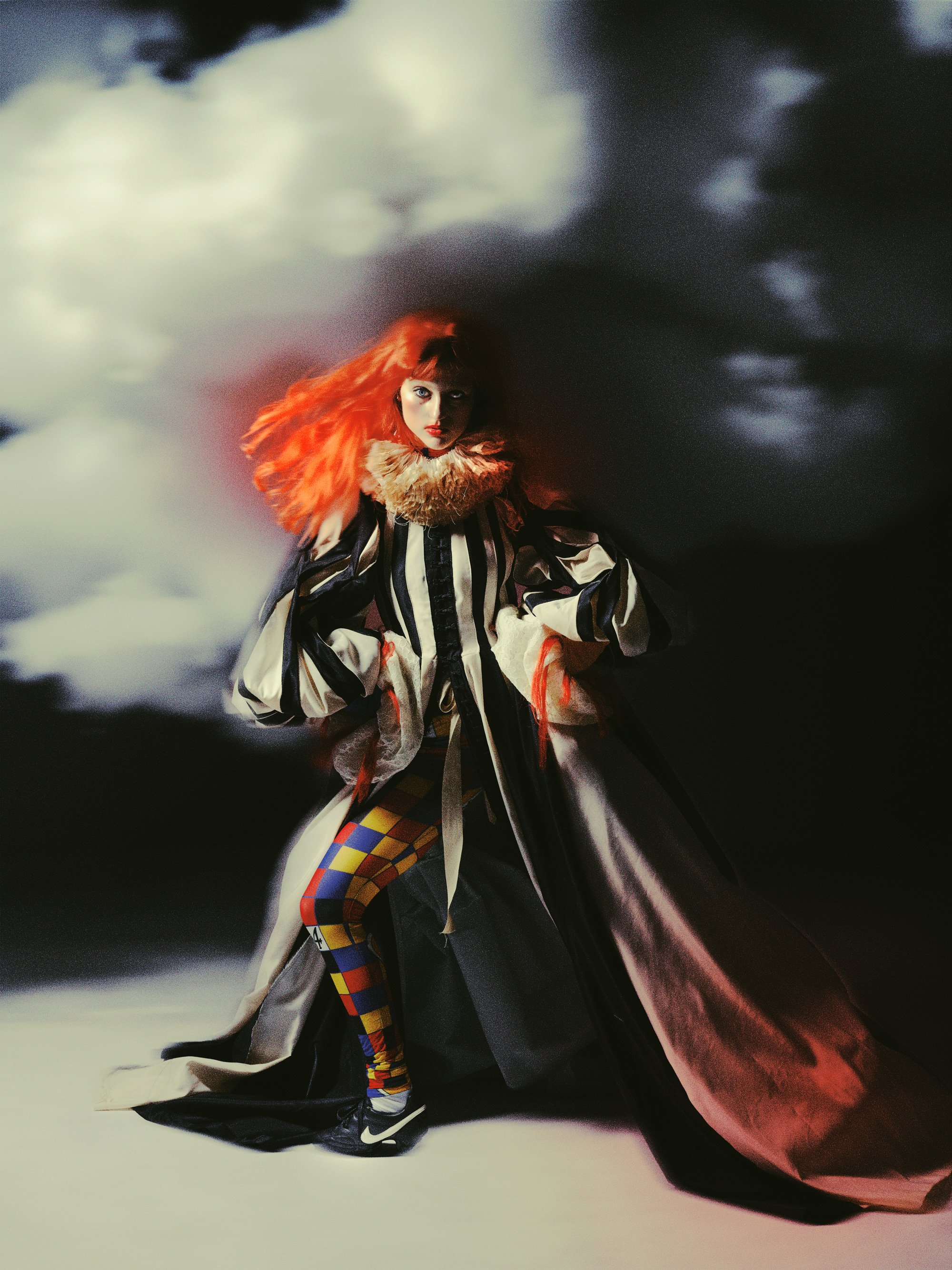 Une femme avec de longs cheveux roux, portant un manteau oversized, un foulard en fourrure et des leggings à motifs colorés, pose devant un fond de ciel nuageux.