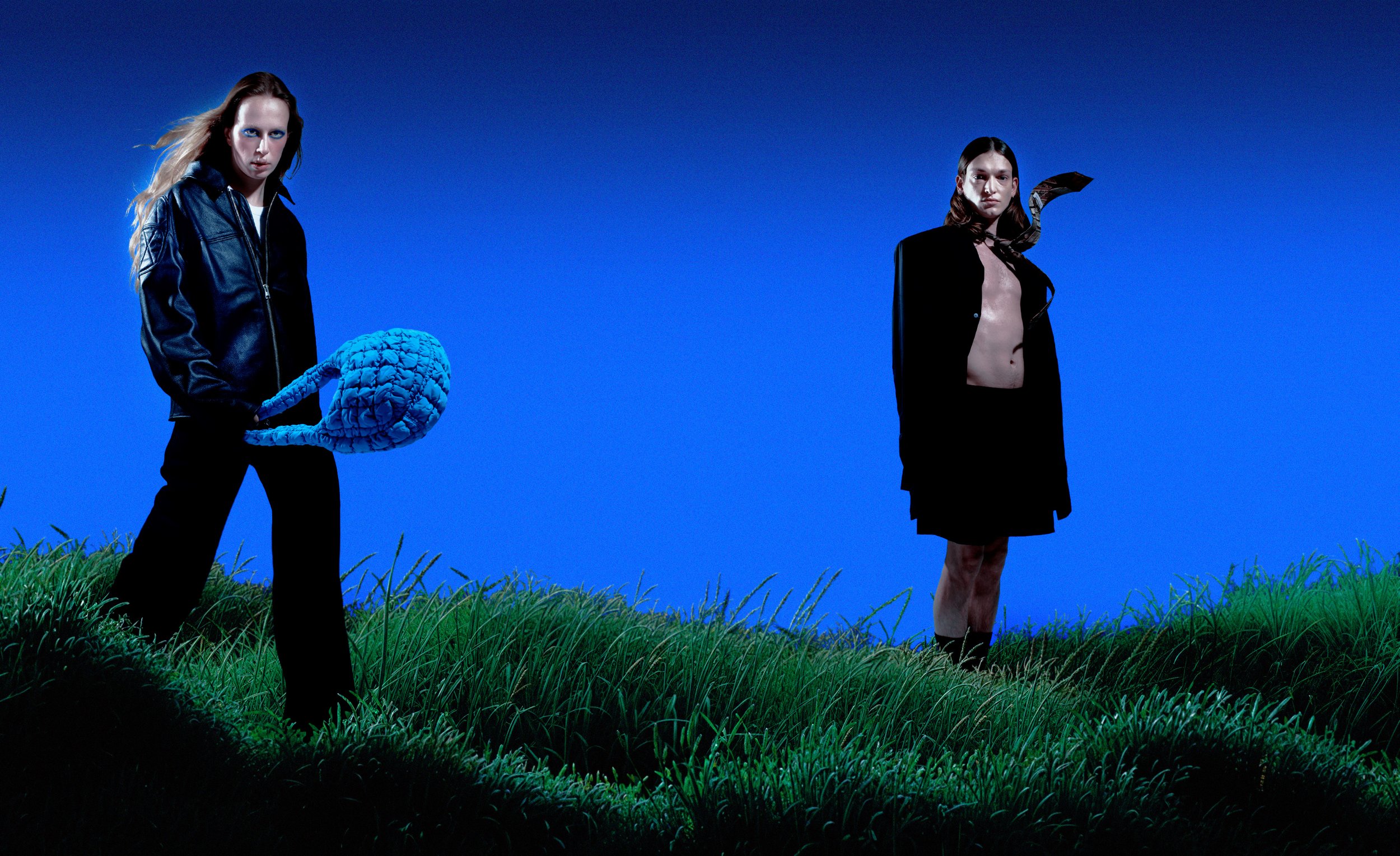 Deux personnes debout dans une prairie verte contre un ciel bleu, une porte-clés en forme d'organe humain dans la main de la personne à gauche, et une petite figure mystérieuse attachée à leur cou