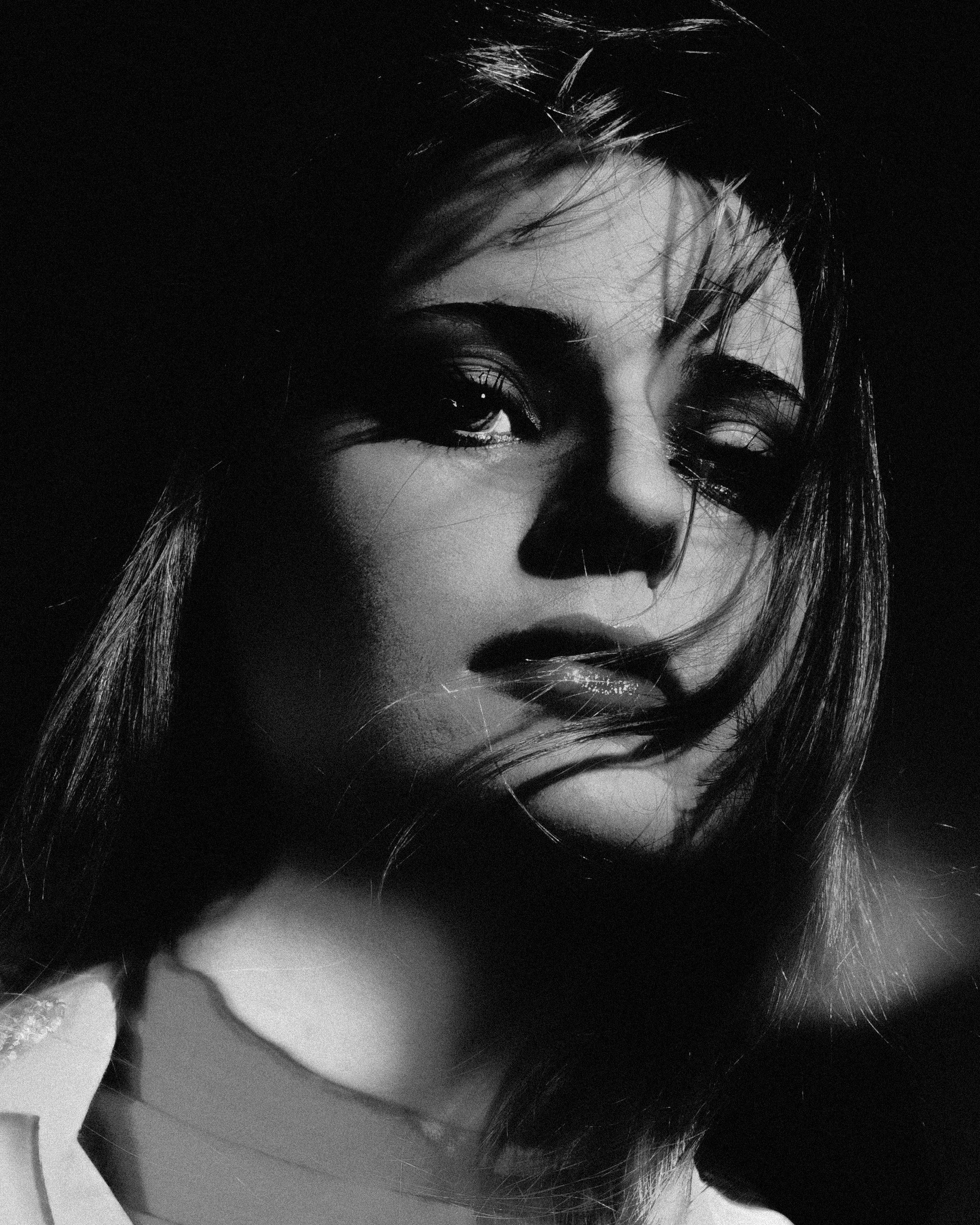 Portrait en noir et blanc d'une femme avec des cheveux longs et sombres, regardant vers la caméra, avec un éclairage dramatique sur son visage.