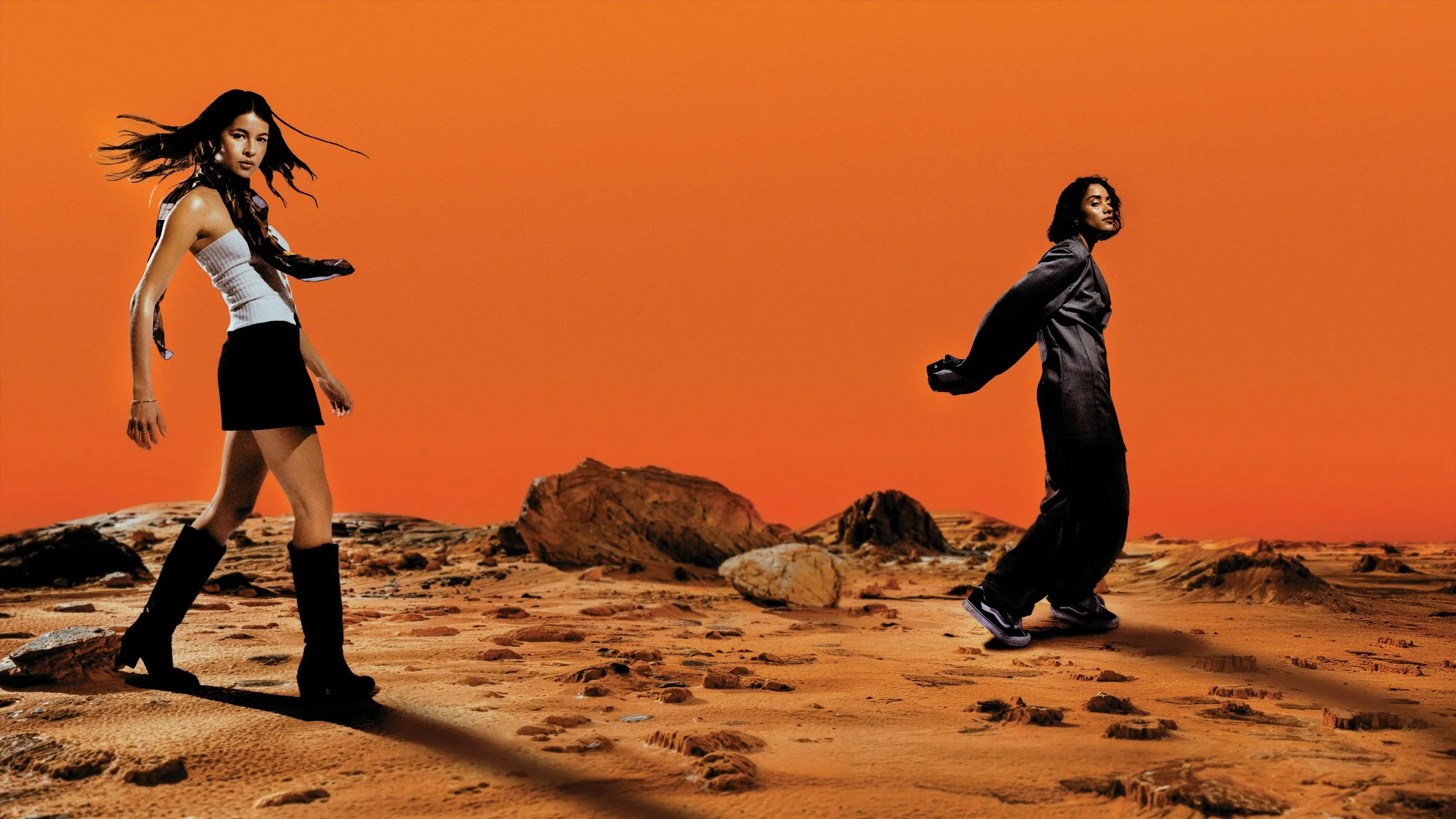 Deux personnes marchant sur un terrain rocheux de couleur orange avec un ciel orange en arrière-plan, l'une vêtue d'une tenue moderne et la autre d'une tenue décontractée.