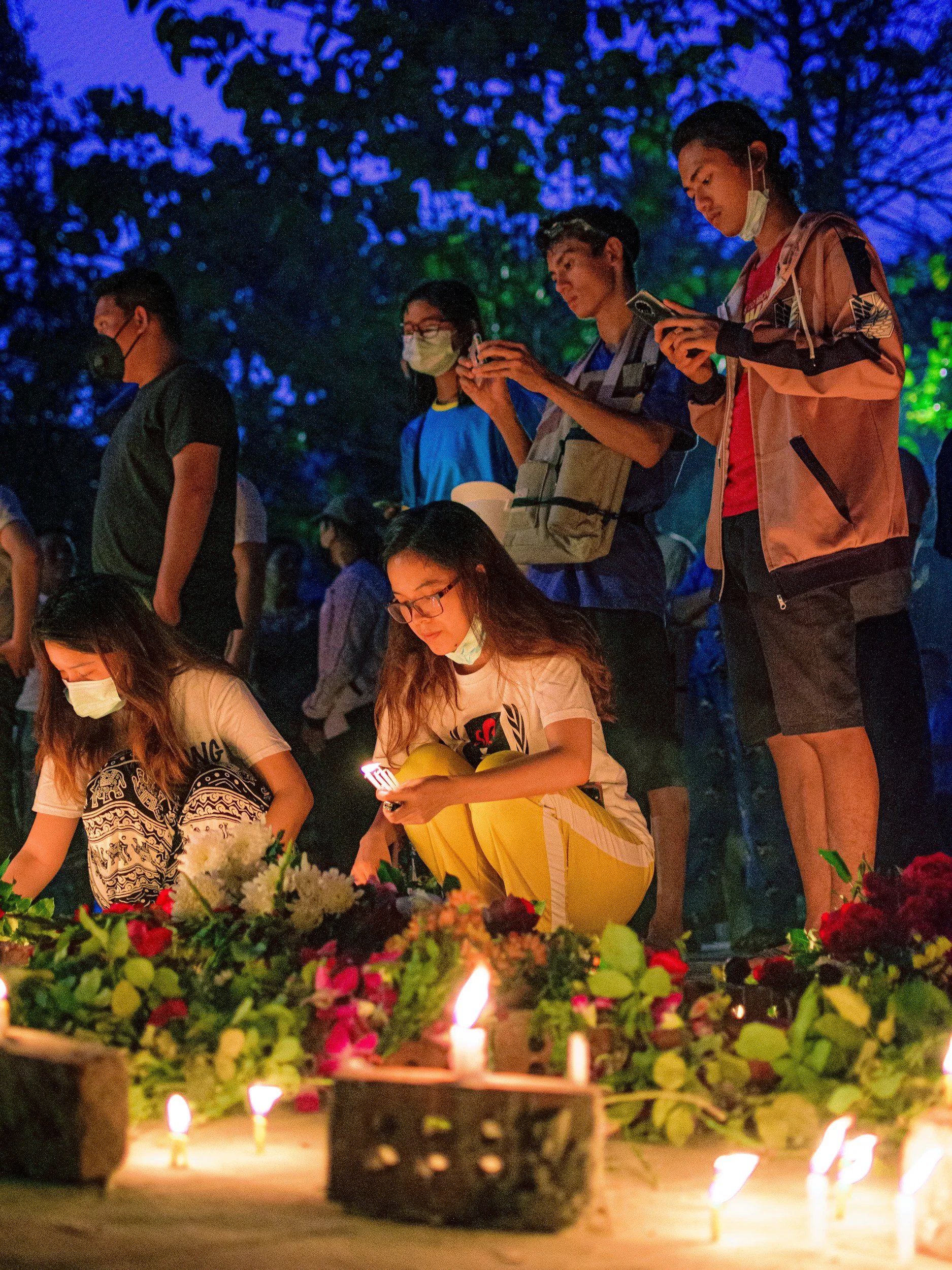 People, including children, participating in a candlelit vigil outdoors at dusk, with some wearing masks and looking at their phones, while others light candles near a floral arrangement.