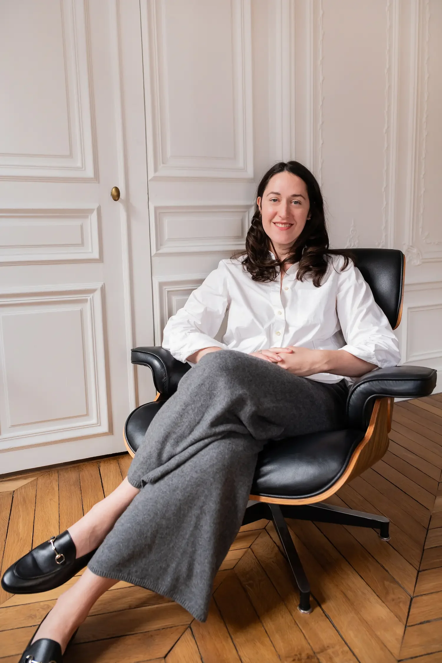 A woman with shoulder-length dark hair, sitting comfortably in a black leather and wood armchair in a room with white paneled walls, wooden flooring, and decorative molding, smiling at the camera.