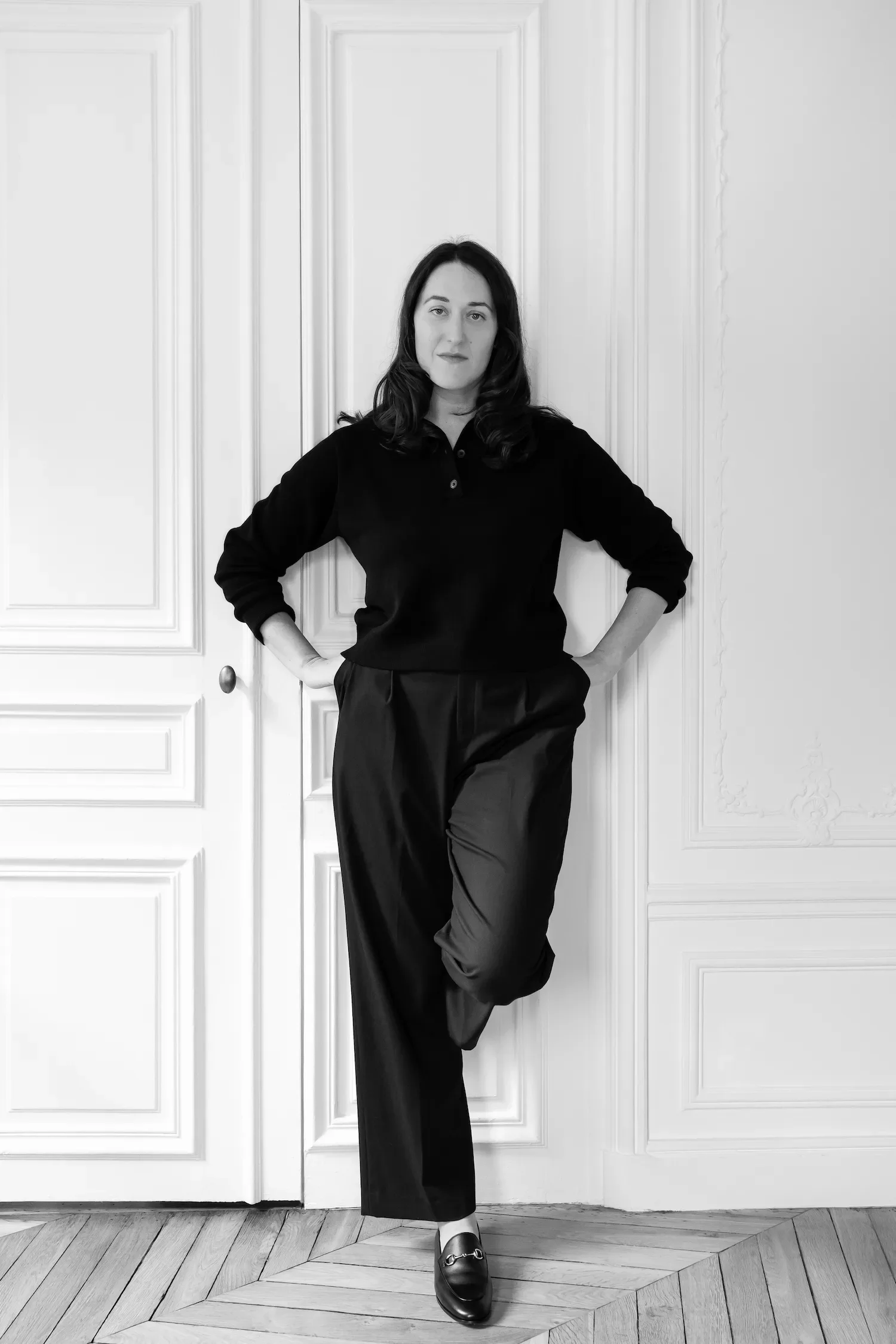 A woman with dark hair, dressed in black, standing against a white paneled wall, with one hand on her hip and her other hand on her waist.