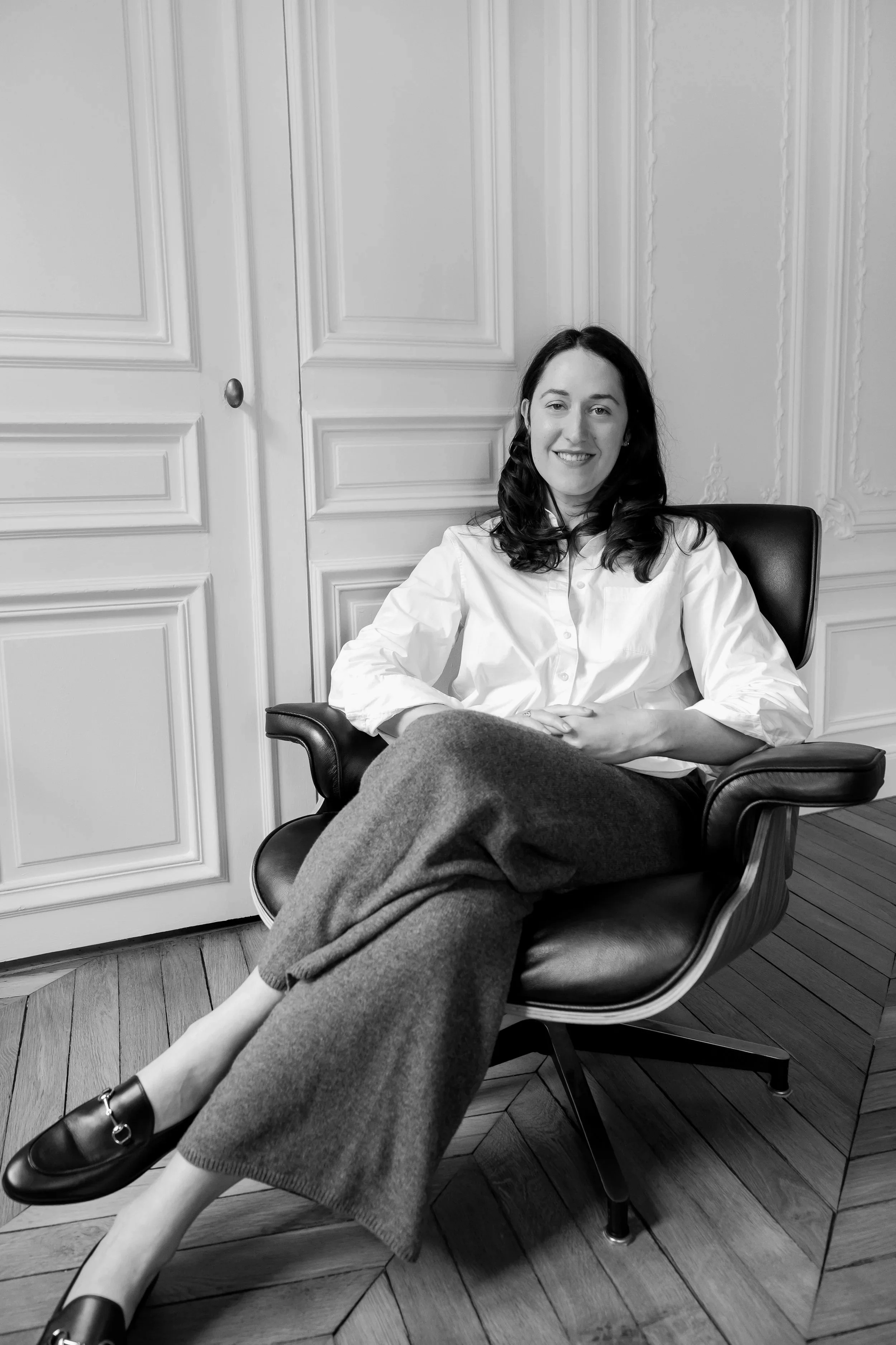 A woman with dark hair wearing a white shirt and dark pants sitting on a black leather chair in a room with paneled walls and wooden flooring.
