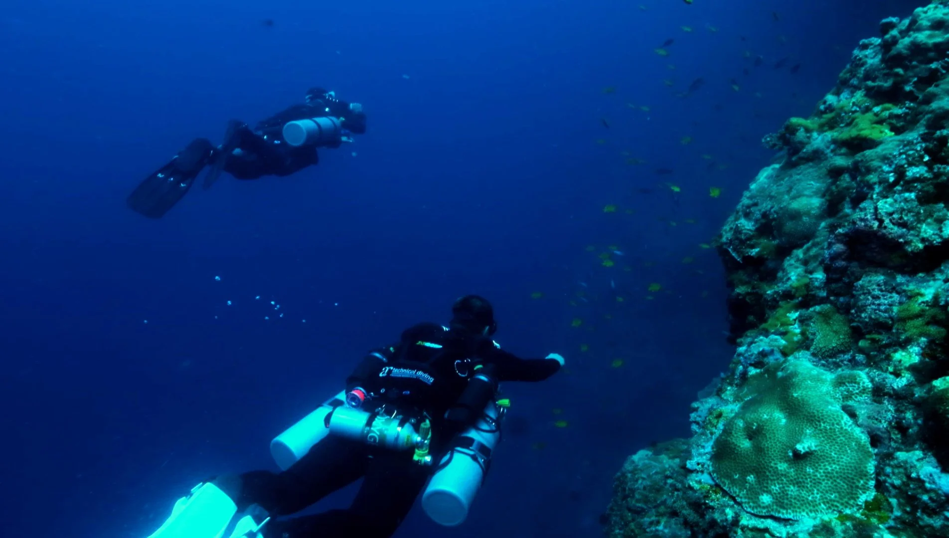 Two scuba divers exploring a coral reef underwater near a rocky wall.