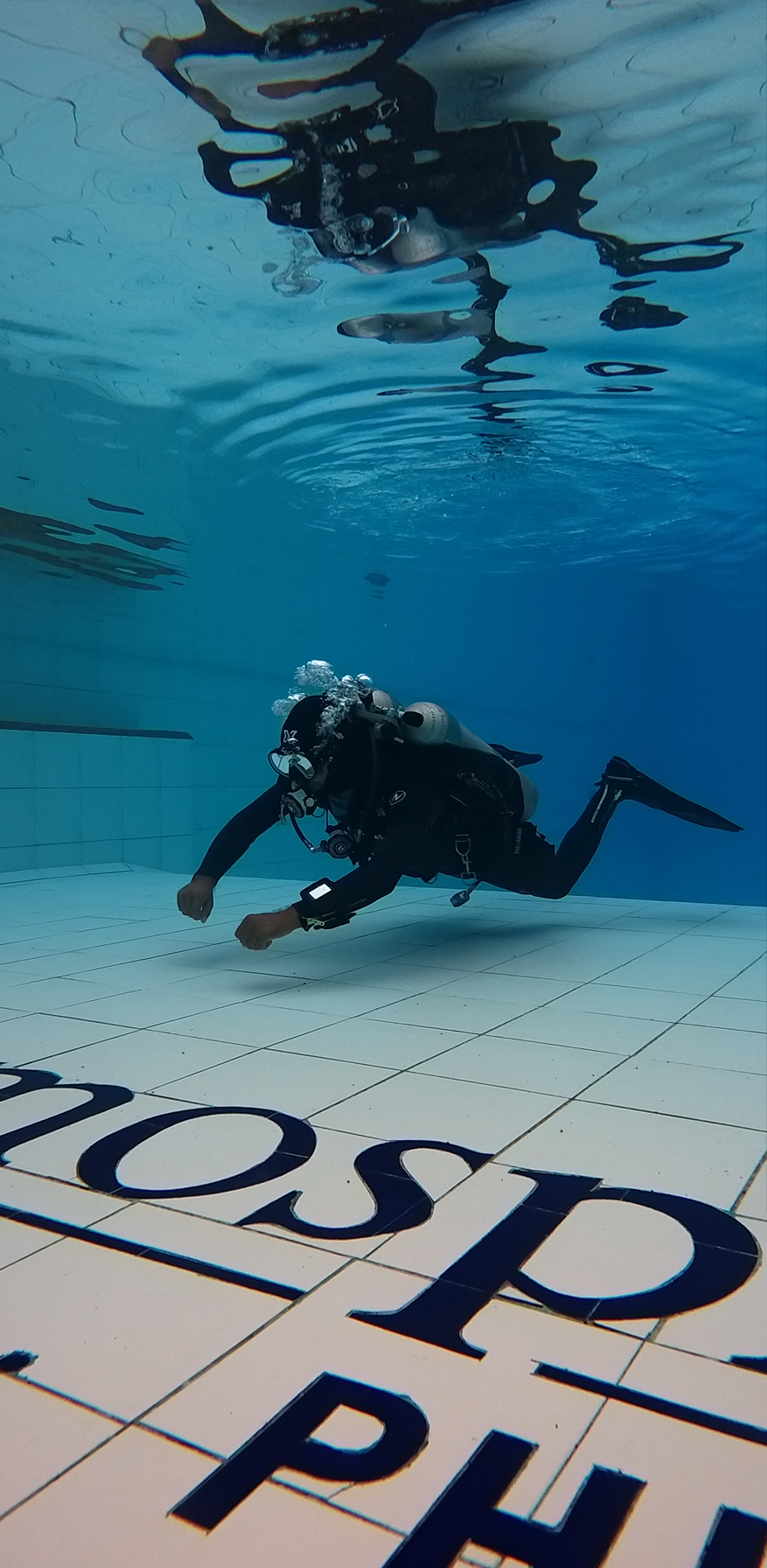 A scuba diver underwater in a swimming pool, wearing a wetsuit, mask, and oxygen tank, exploring the pool's bottom near the tiles.