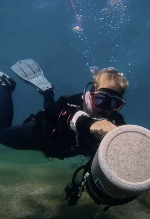 Person in scuba gear underwater holding a large, round object, with a small boat and water in the background.