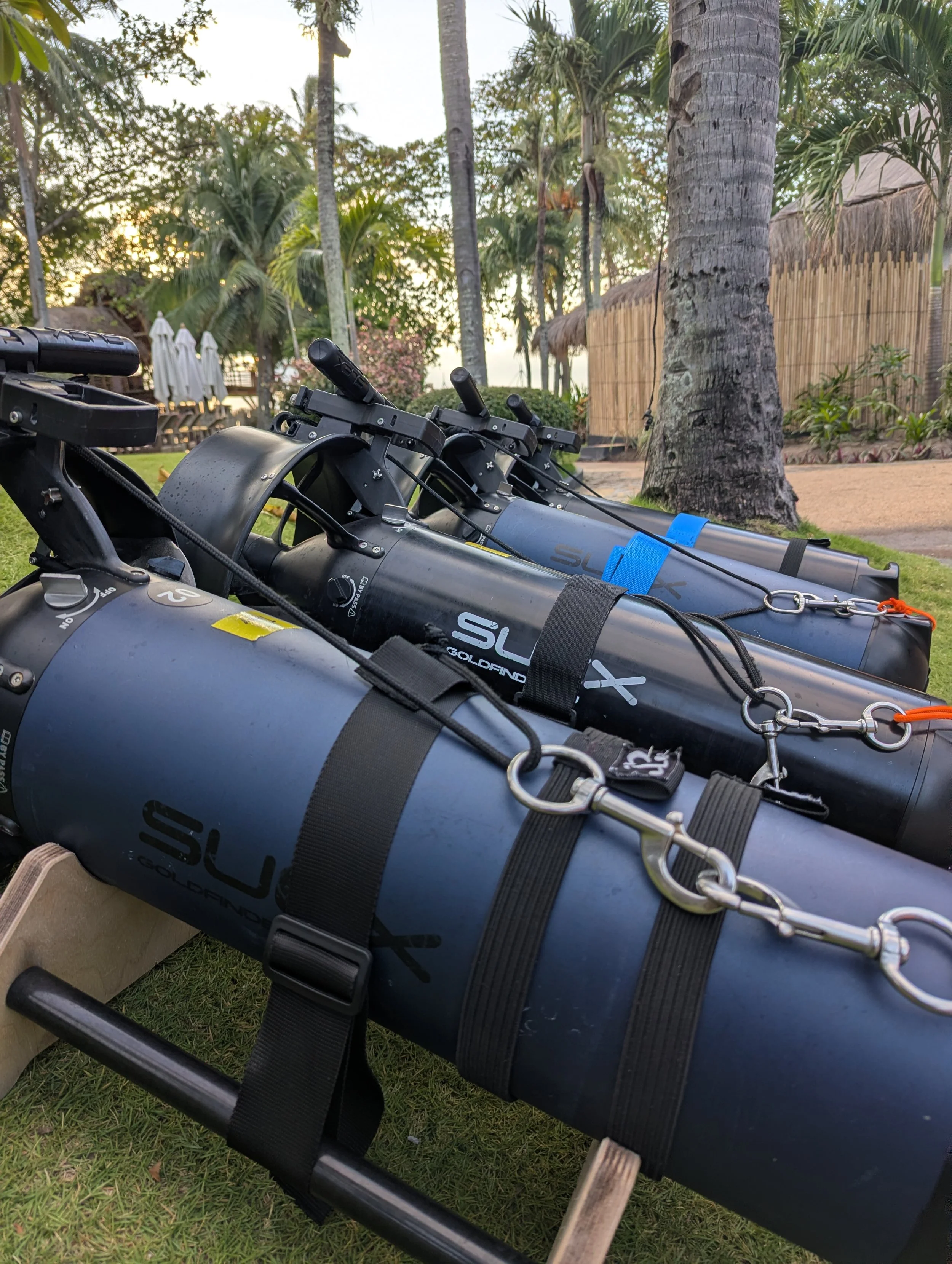 Snorkel fins with black bands attached, laid on grass near a wooden block, trees, and a bamboo fence in a tropical outdoor setting during sunset.
