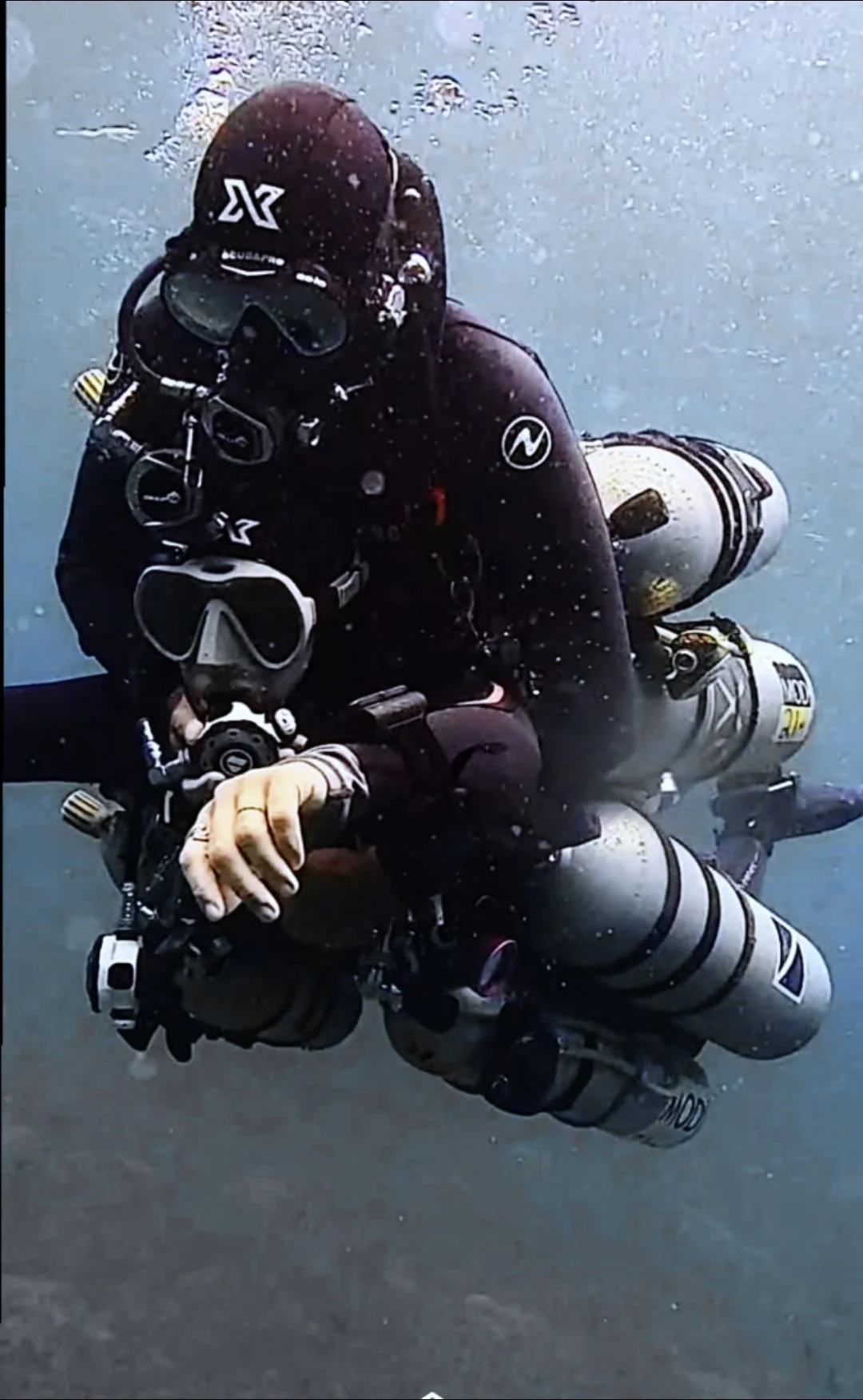 A scuba diver underwater in full gear, including a wetsuit, mask, regulator, and tank, holding onto a camera or equipment.