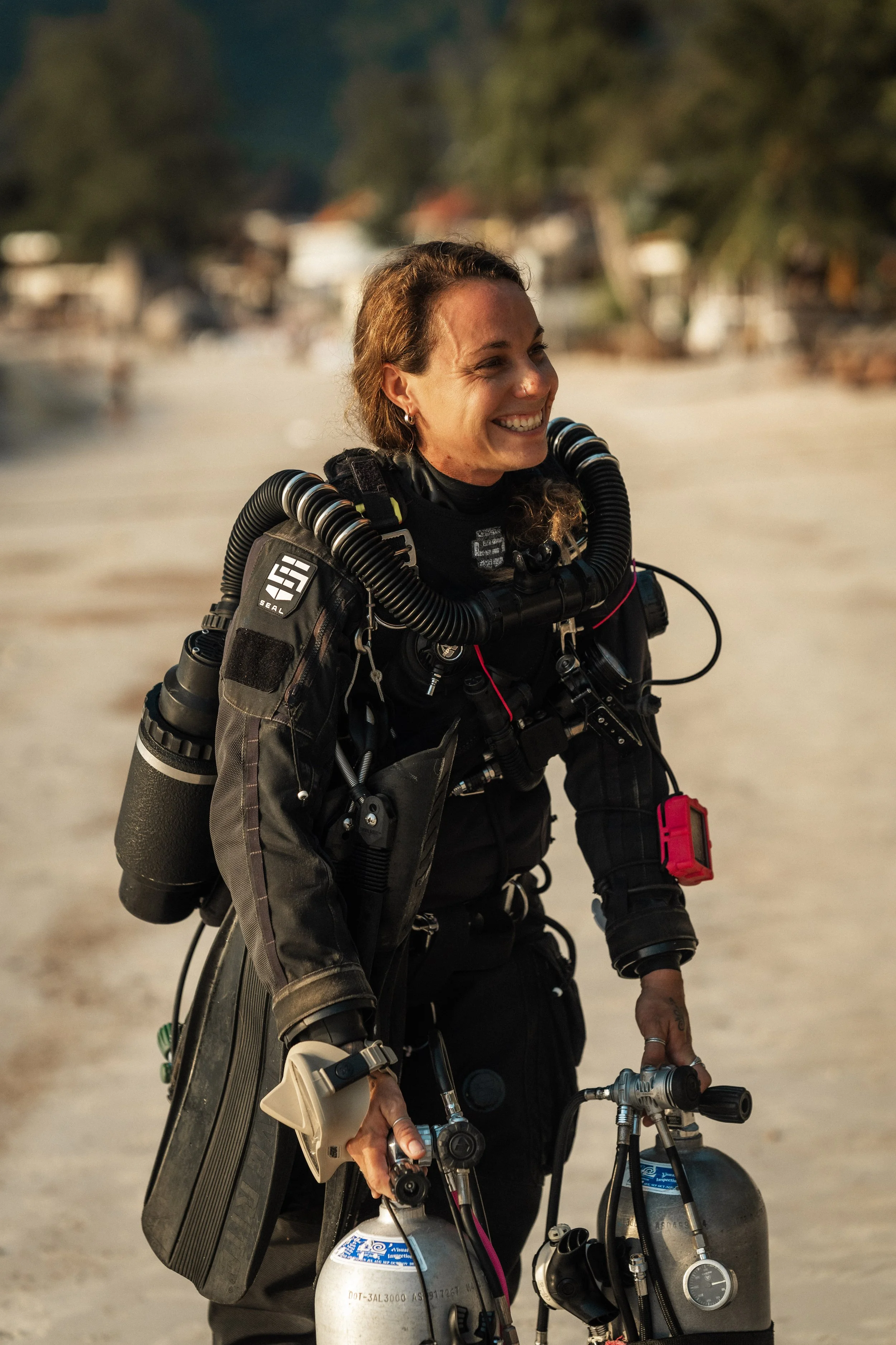 female rebreather diver holding sidemount tanks, smiling on the beach