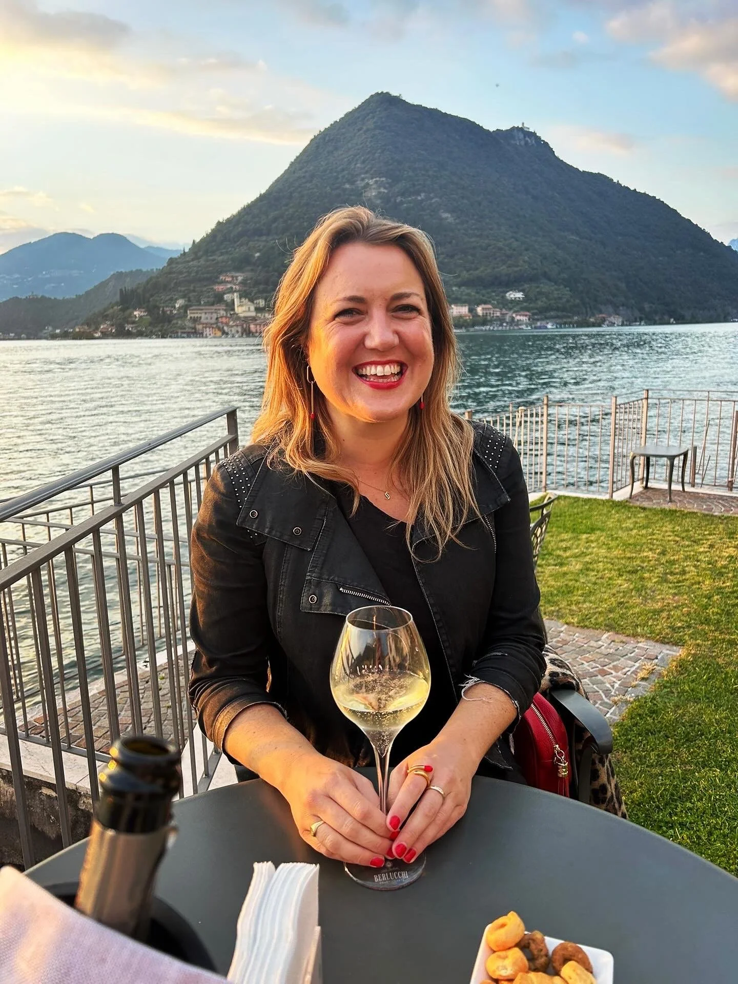 Sophie McLean in Franciacorta at an outdoor table by lake Iseo, holding a glass of sparkling wine with mountains in the background.