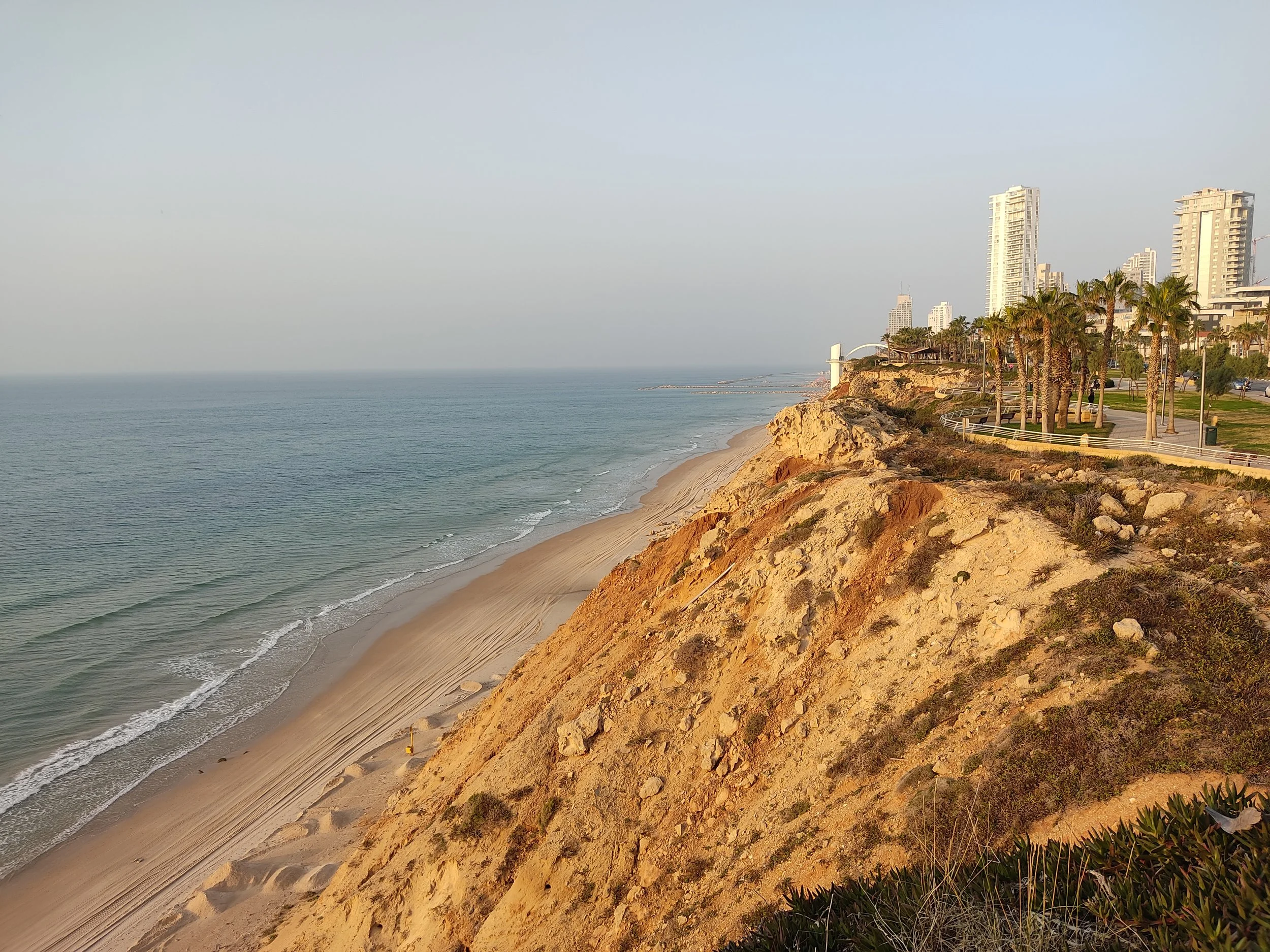 View of a sandy beach in Netanya with tall buildings, palm trees, and a rocky cliff during sunset.