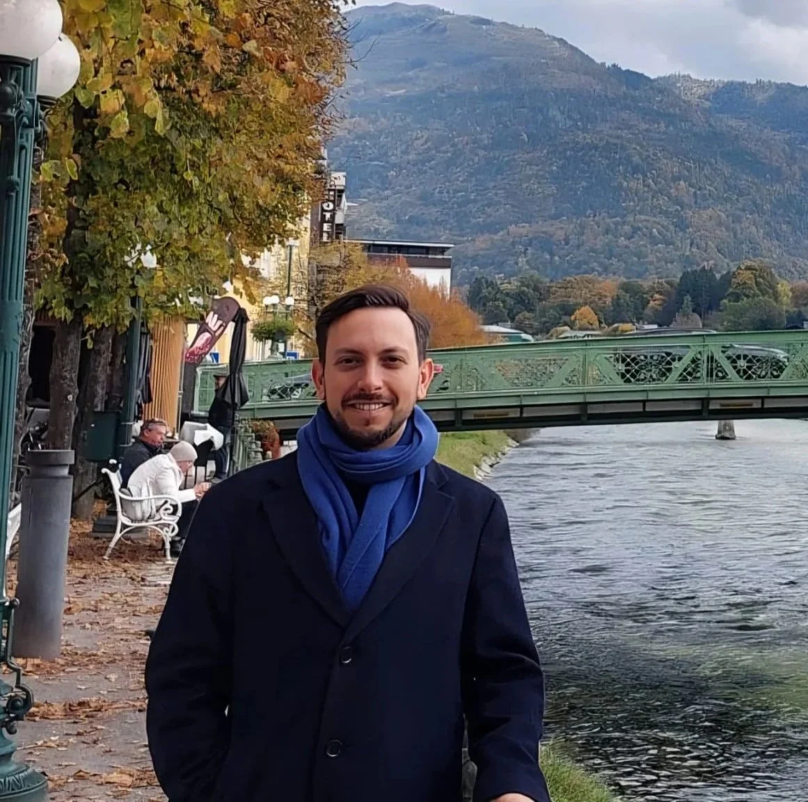 Idan Levy, the site owner, in a black coat and blue scarf smiling while standing next to a river with a green bridge overhead. Behind him, there are trees with autumn foliage and mountains in the background.