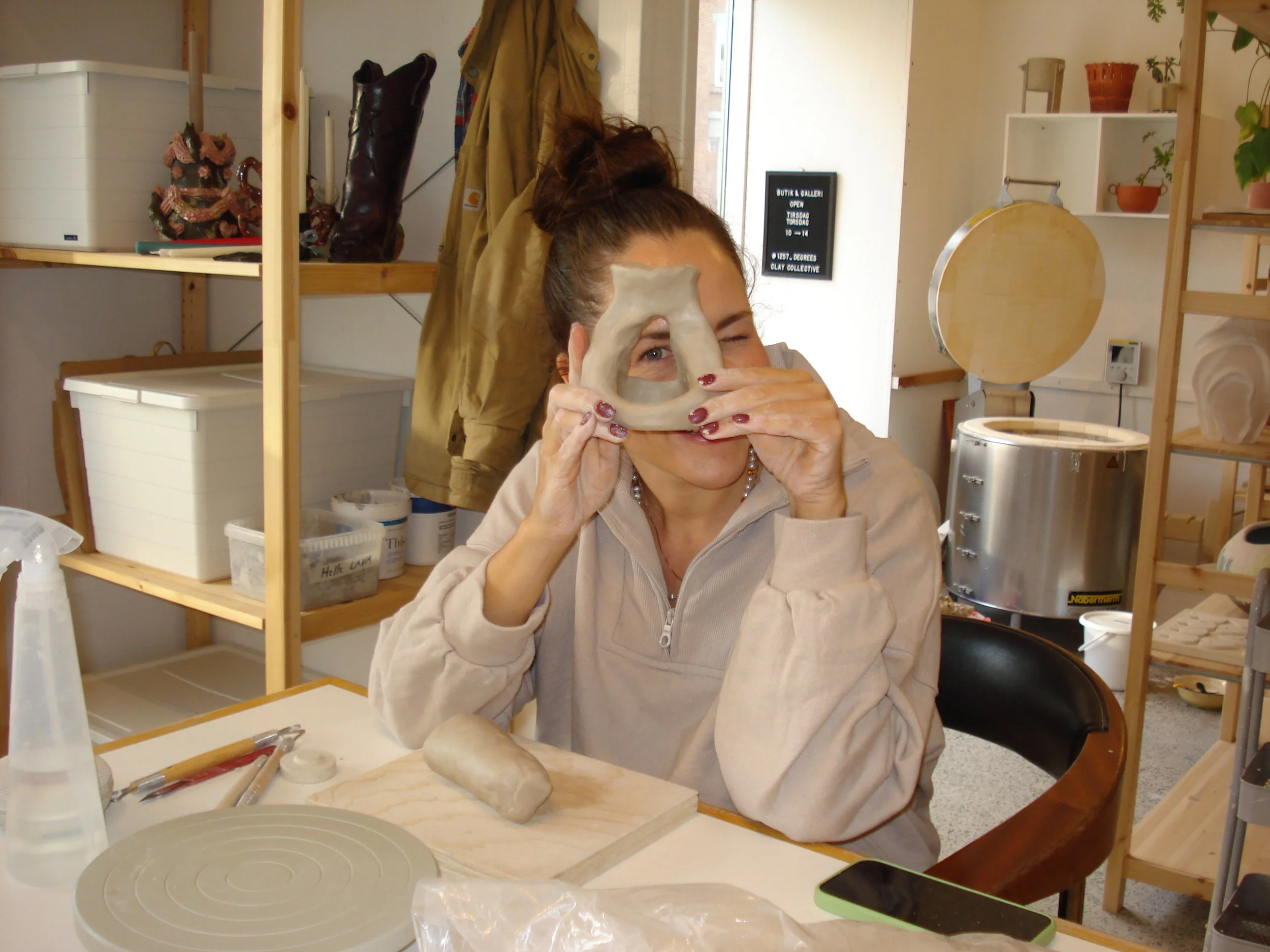A woman with her hair in a bun holding a clay or ceramic object to her face, winking. She is sitting at a workspace with pottery tools, a spray bottle, a cylindrical clay piece on a wooden board, and a smartphone on the table. The background shows wo