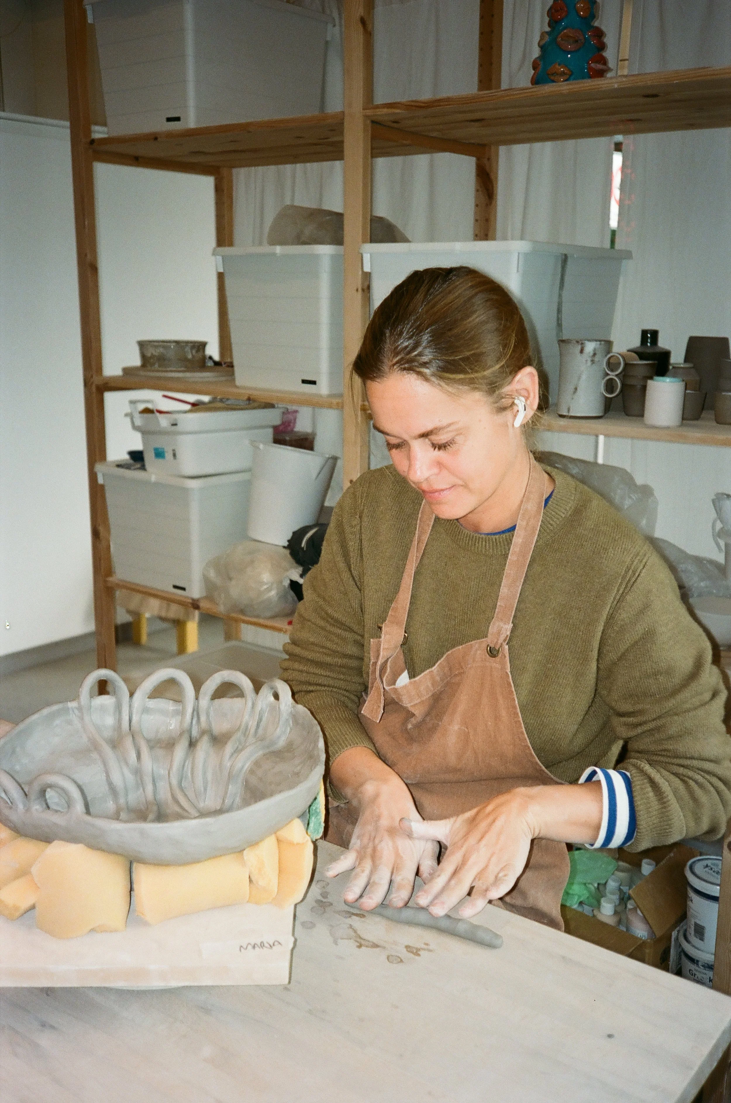 A woman working on pottery in a ceramics studio, wearing a brown apron, with shelves of ceramic items in the background.