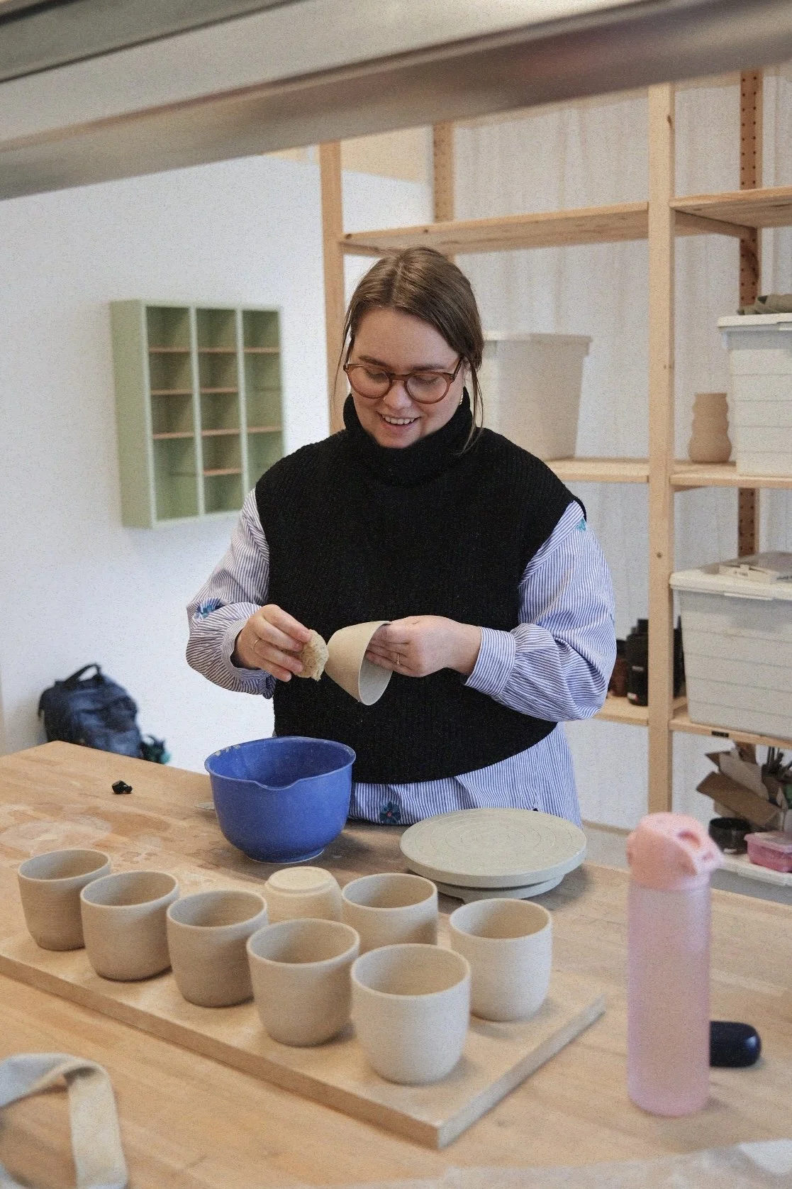 A woman with glasses and a black vest is decorating small handmade ceramic cups on a wooden table. There are multiple unglazed cups, a blue bowl, and a pink bottle on the table. She is smiling while working in a pottery studio.