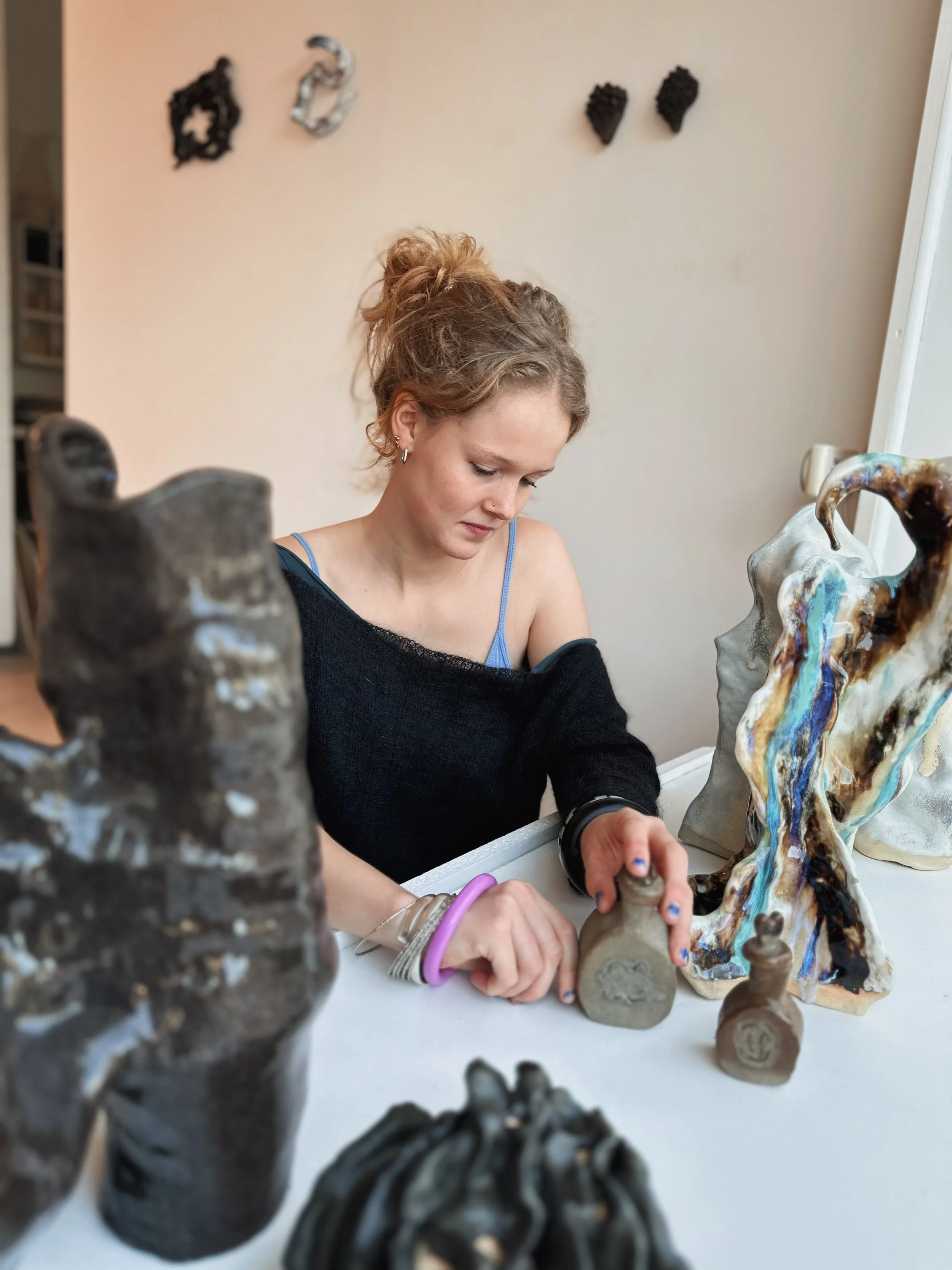 A woman with red curly hair working on clay sculptures at a table, with colorful and black abstract sculptures around her.