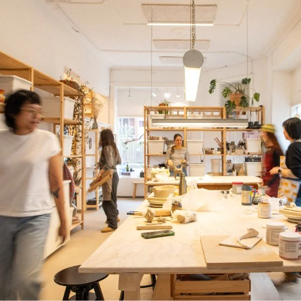People moving and working in a pottery studio in Copenhagen with shelves of clays and ceramics, and a large table with pottery materials.