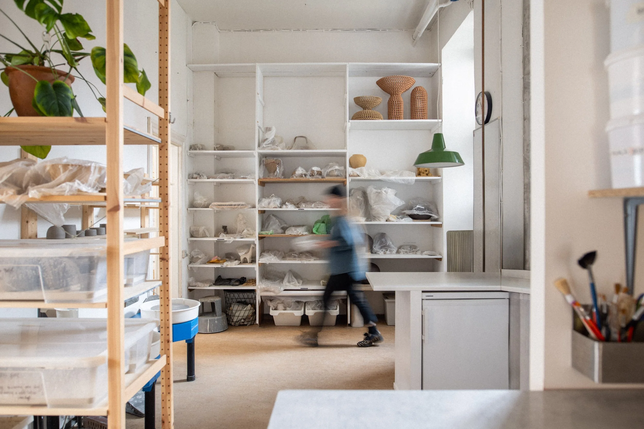 Interior of a ceramics workshop with white shelving units filled with various clay and ceramic items, potted plants and a person walking past. 