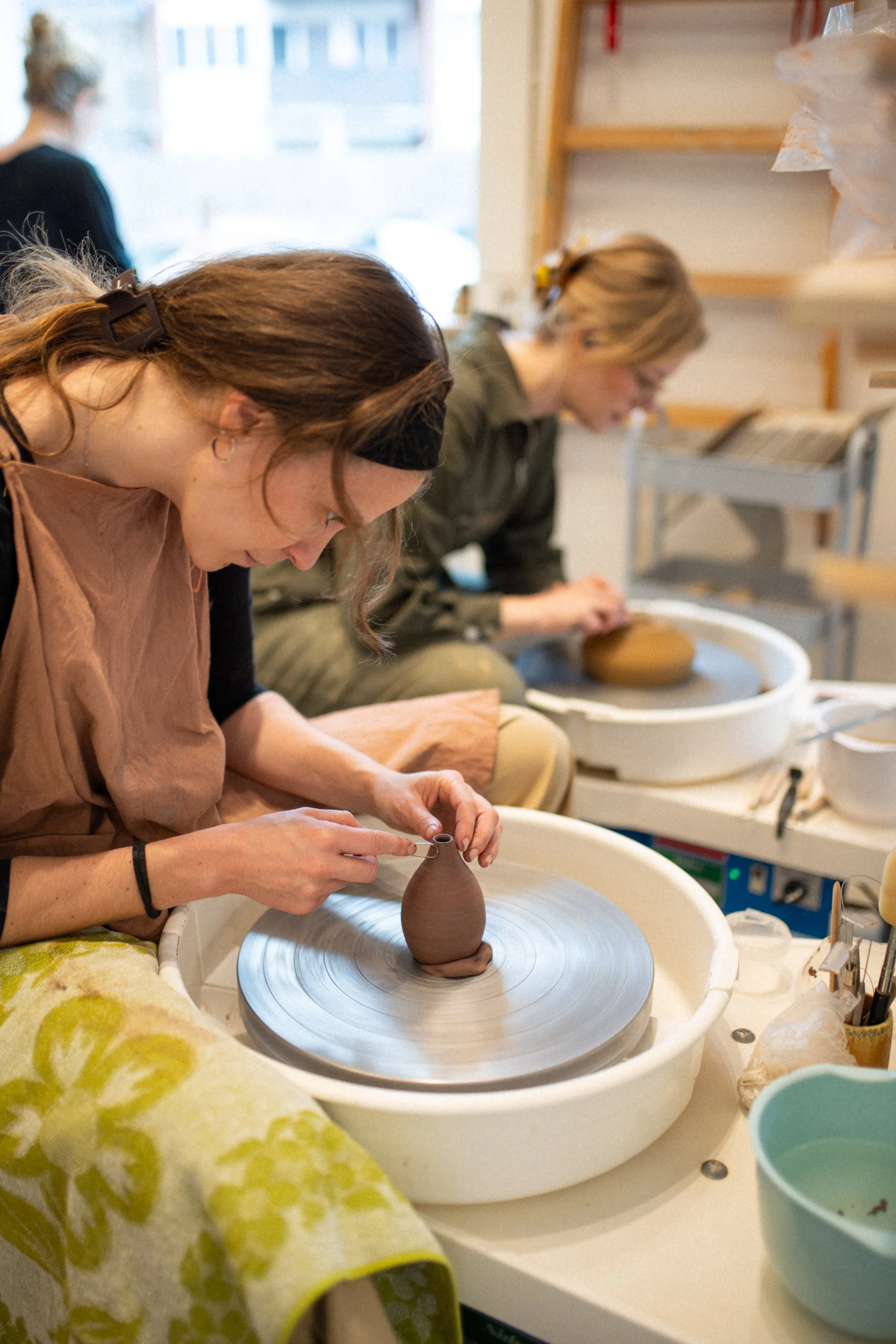 Two women creating a ceramic pot on a pottery wheel while working in a pottery studio.