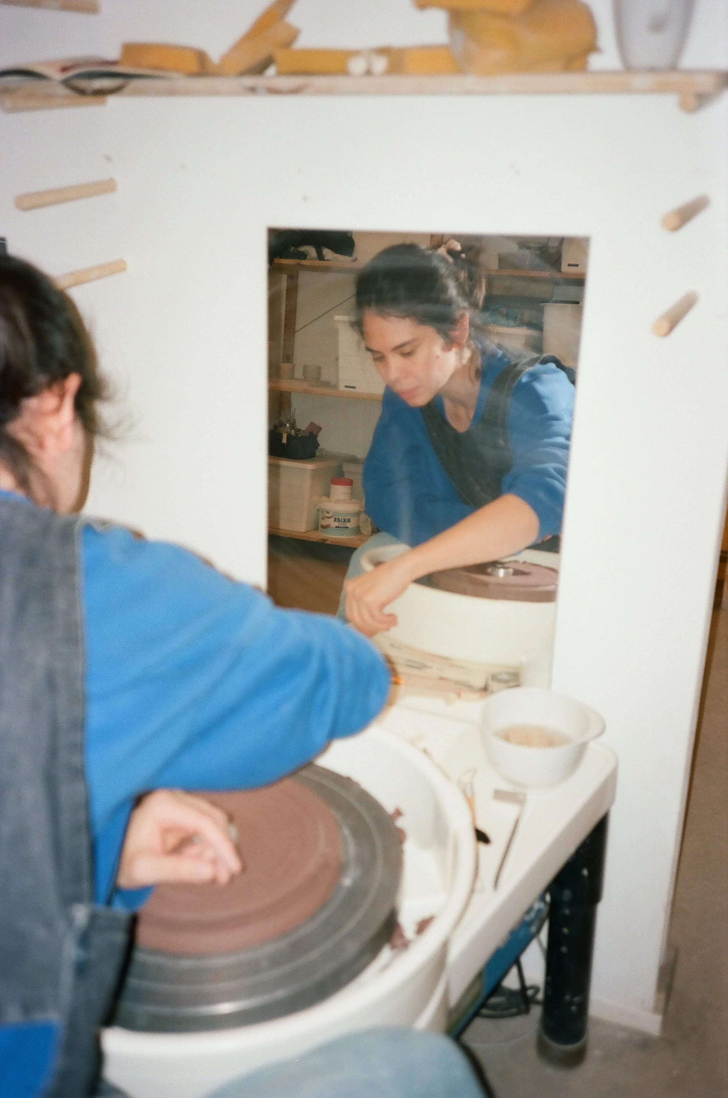 A woman working on a pottery wheel, seen through a mirror, with her reflection visible in the mirror behind a white frame.