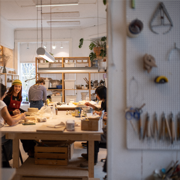 People working in a ceramics or pottery studio, where they are shaping clay on tables and where there are shelves holding supplies and tools in the background.