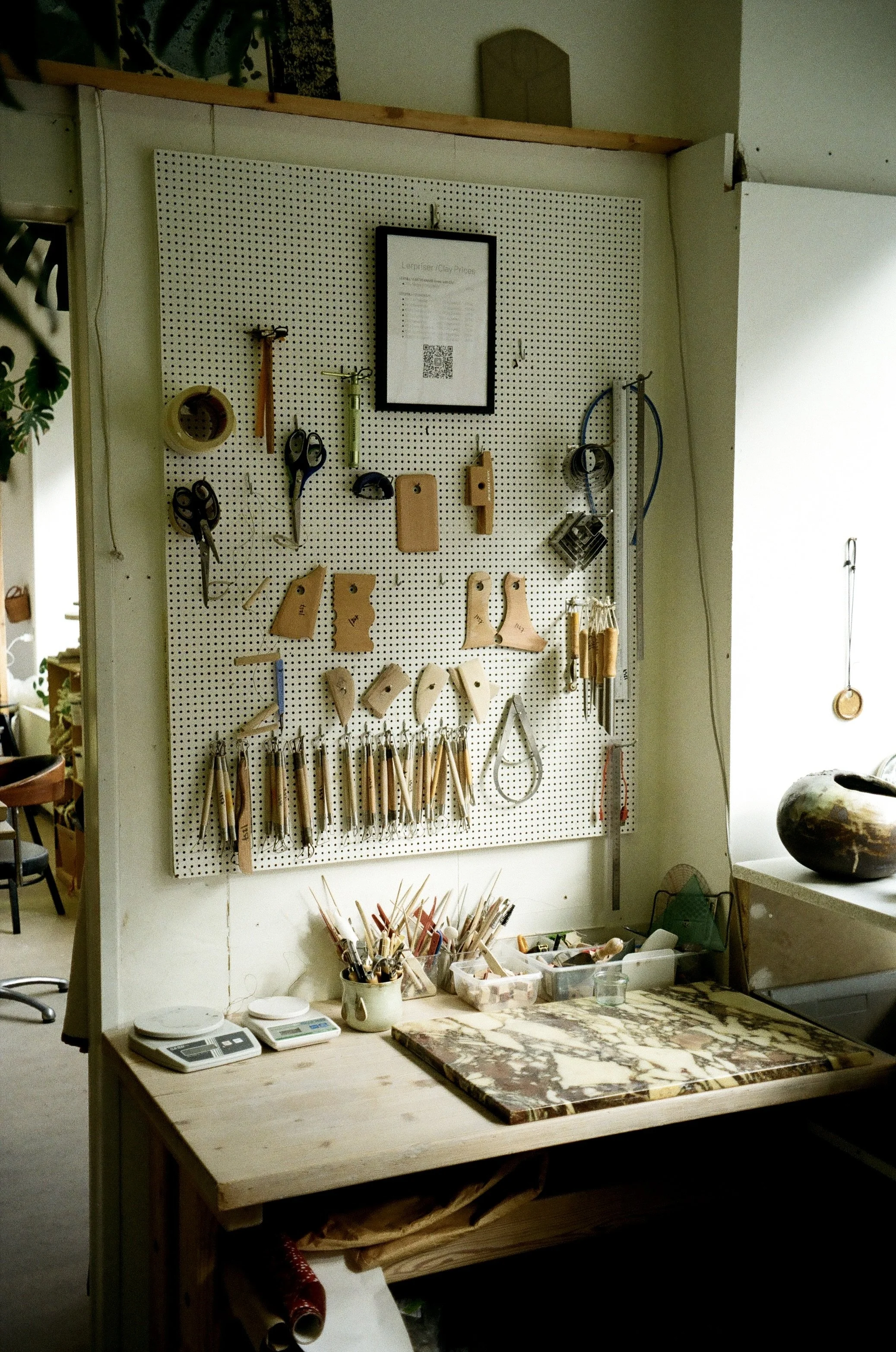 A woodworking workshop with a pegboard holding tools like scissors, knives, and measuring tapes. There is a framed price list hanging on the board. The wooden workbench below has various art supplies, a digital scale, and a marble slab. A large decorative pot is on the right on a windowsill.