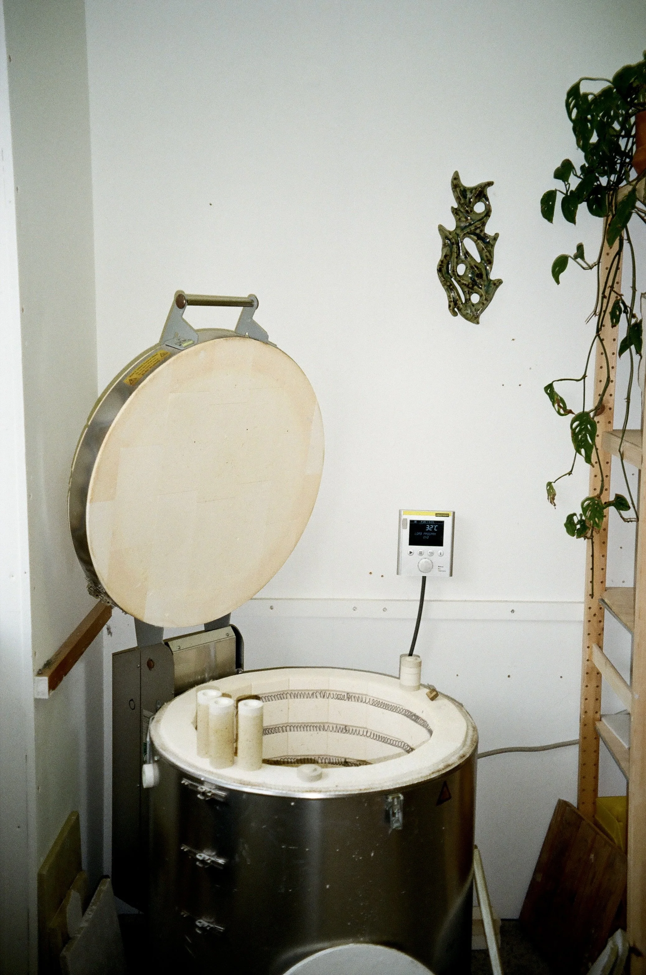 An ceramic kiln with its top open, placed in a room with a digital control panel on the wall, decorated with a metal wall art and a plant on a shelf.