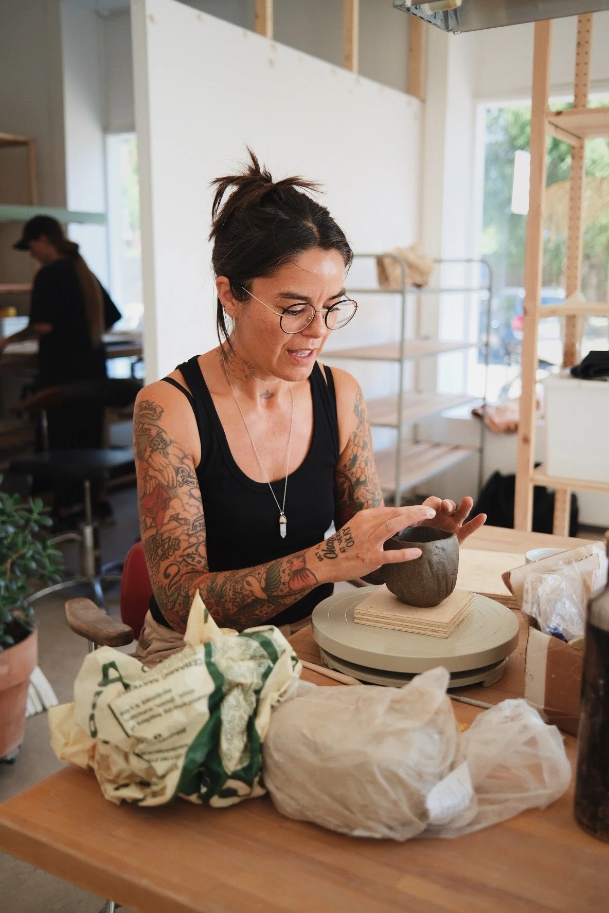 A woman with tattoos and glasses working on clay pottery in a studio, with pottery supplies and a plant on the table.