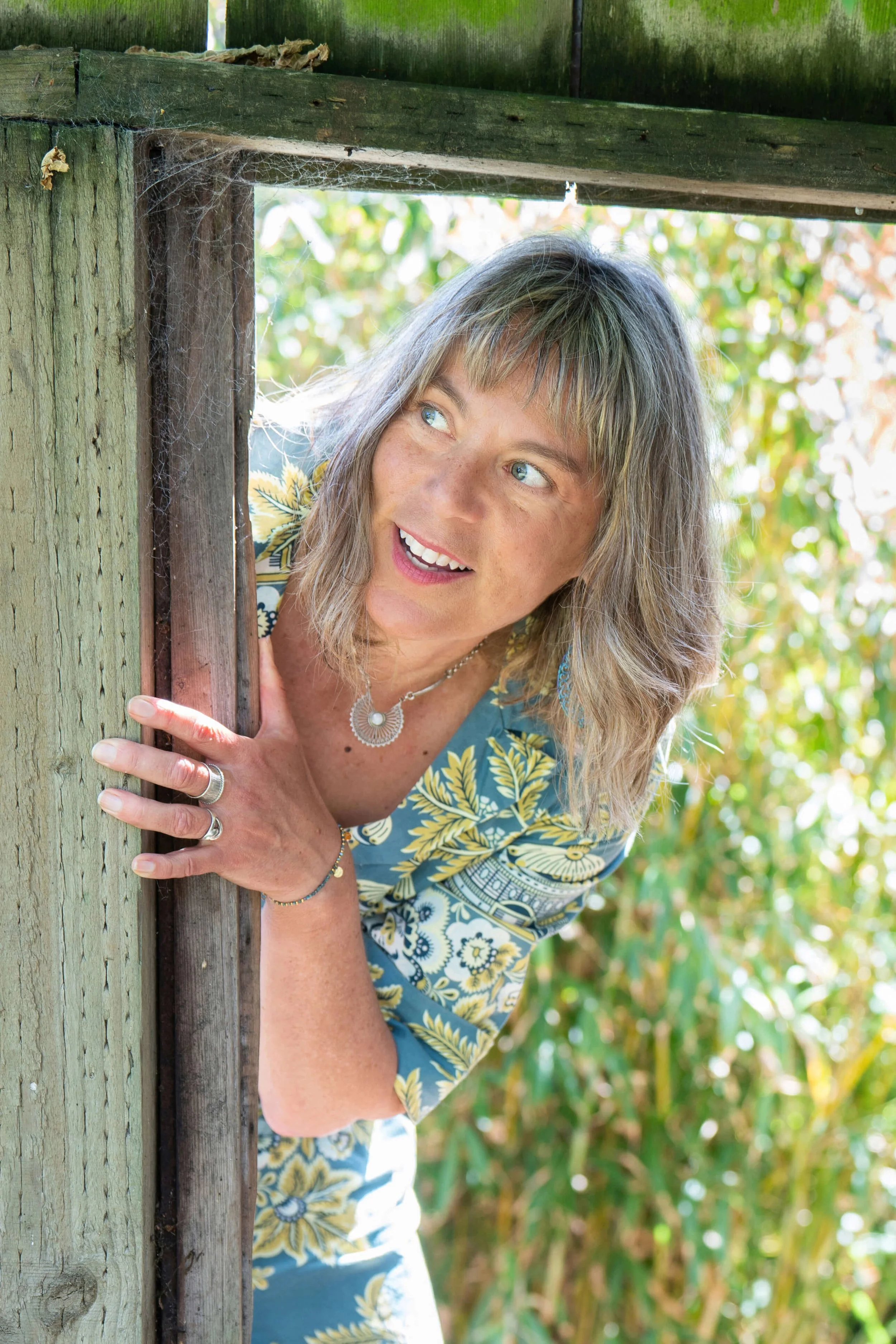 Dashka Slater smiling and peeking out from a wooden structure in an outdoor garden setting.